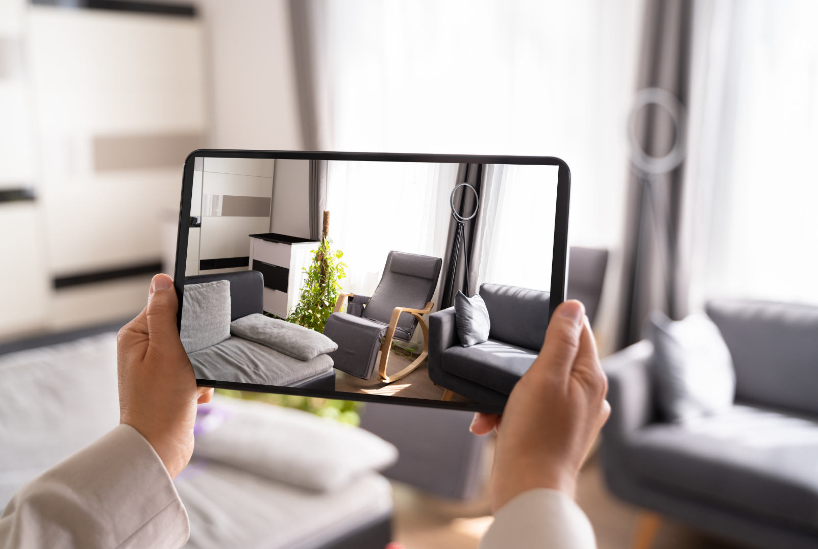 Person taking a photo of a living room with a smartphone, featuring a gray armchair, a black sofa, some cushions, a plant, and a tall group light near the window.