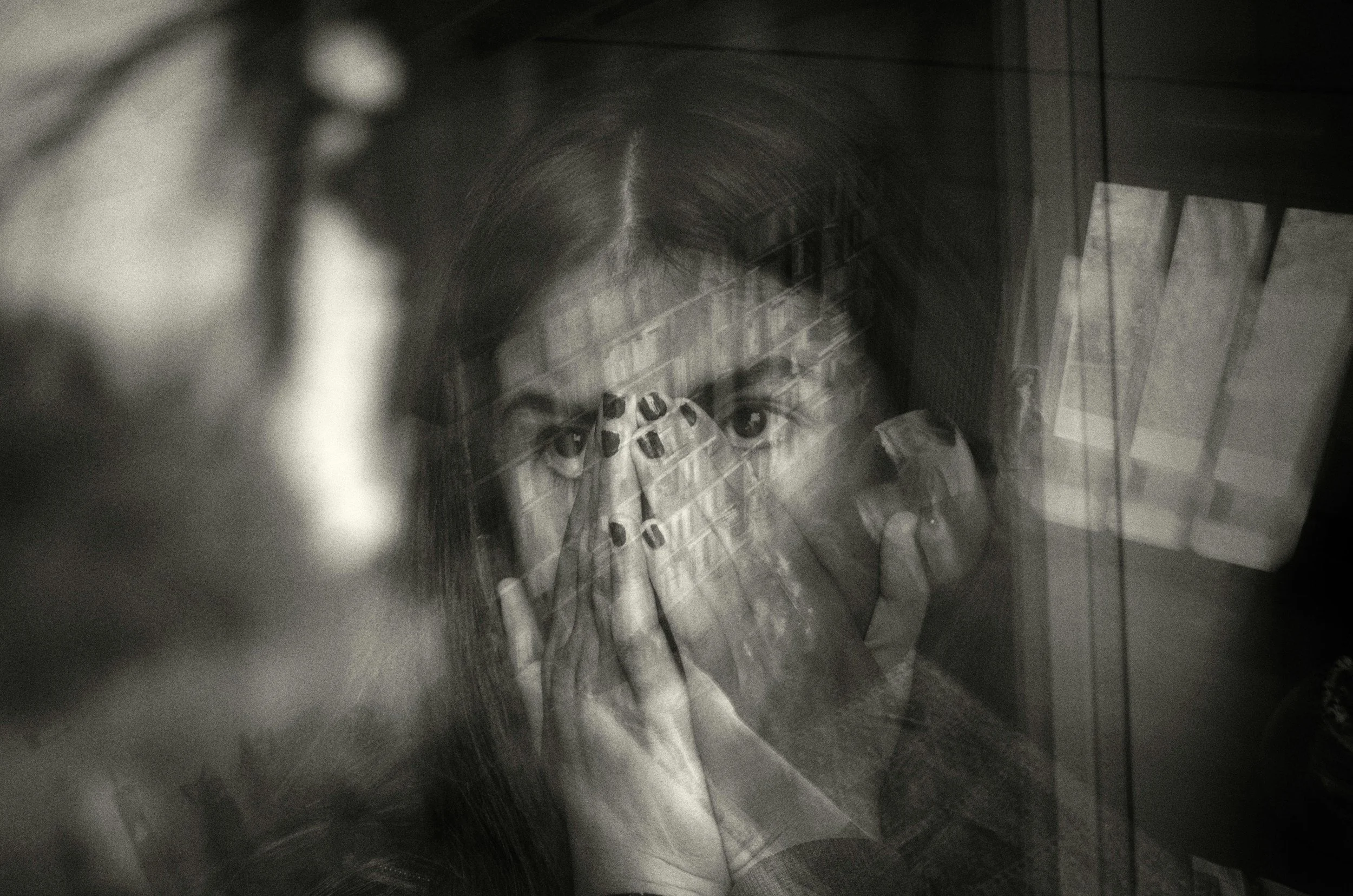 A person with long hair looking through a window with reflections of a staircase, hands with painted nails partially covering their face, creating a layered, surreal effect.