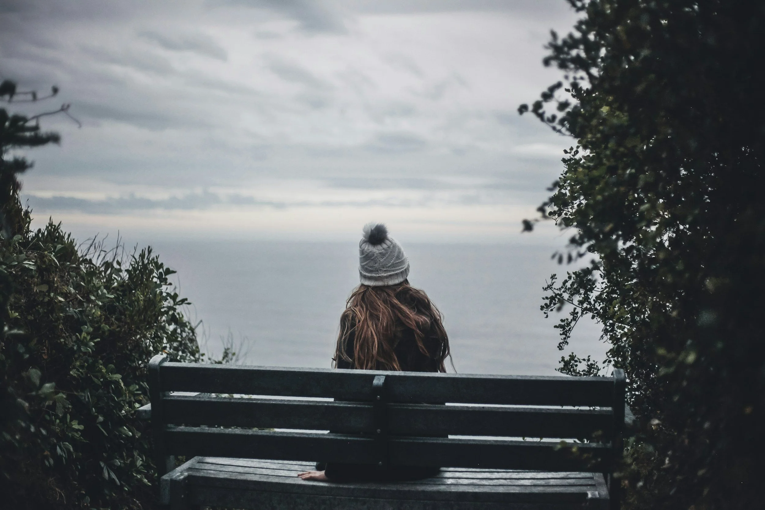A woman sitting on a park bench facing the ocean, wearing a beanie and surrounded by bushes, with overcast sky.
