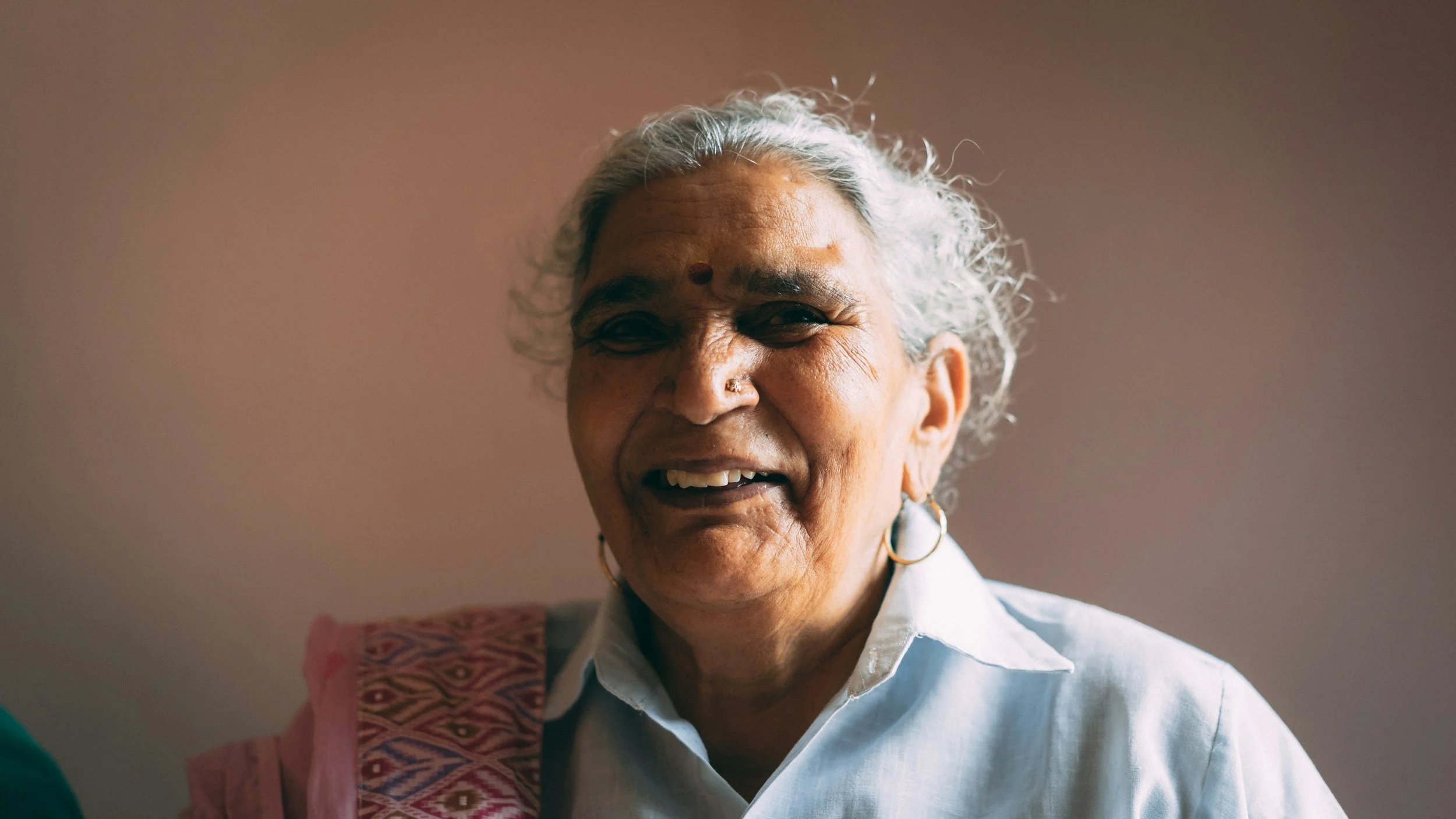 Smiling elderly woman with gray hair, wearing a white shirt and gold earrings, against a plain wall.