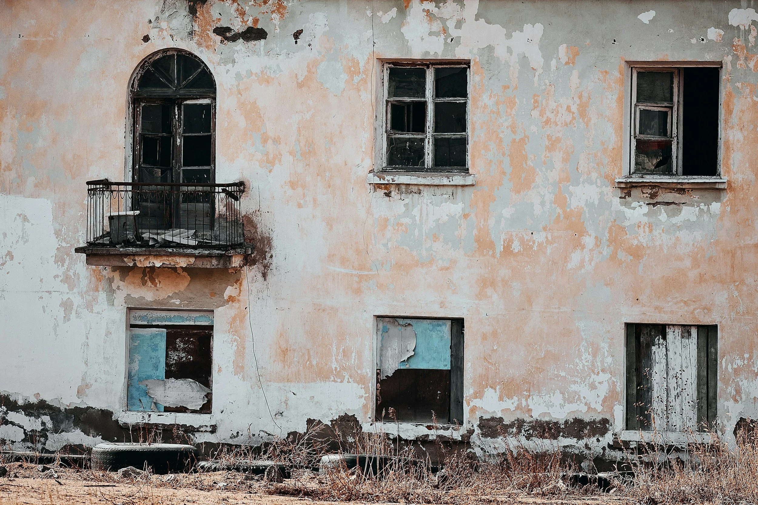 An abandoned building with peeling paint, broken windows, and overgrown weeds at the base.