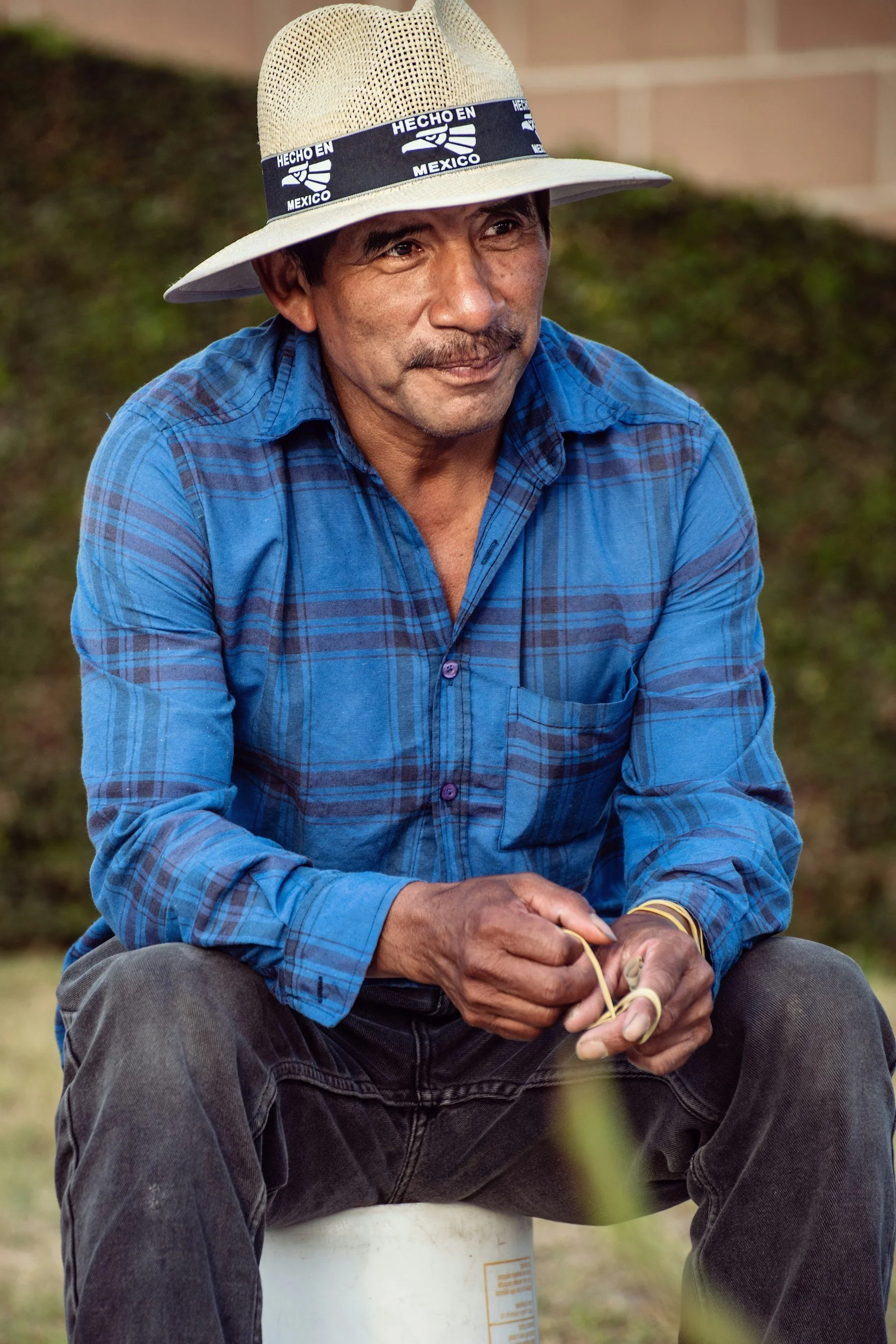 A man with a mustache and dark hair wearing a blue plaid shirt and a wide-brimmed beige hat, sitting outdoors on a white bucket, holding a string or cord, with a blurred natural background.