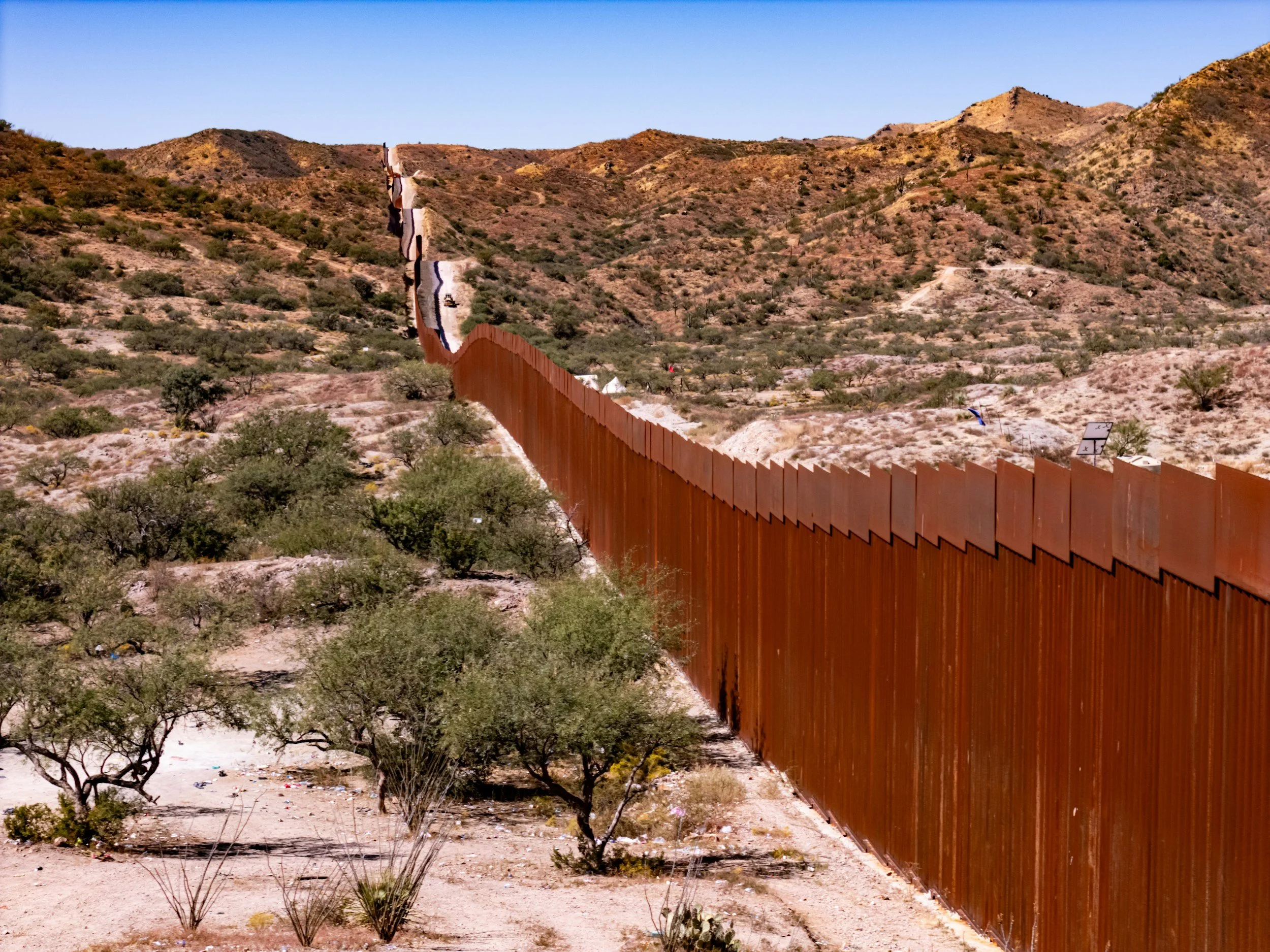 A long border fence running through a desert landscape with hills and sparse vegetation.