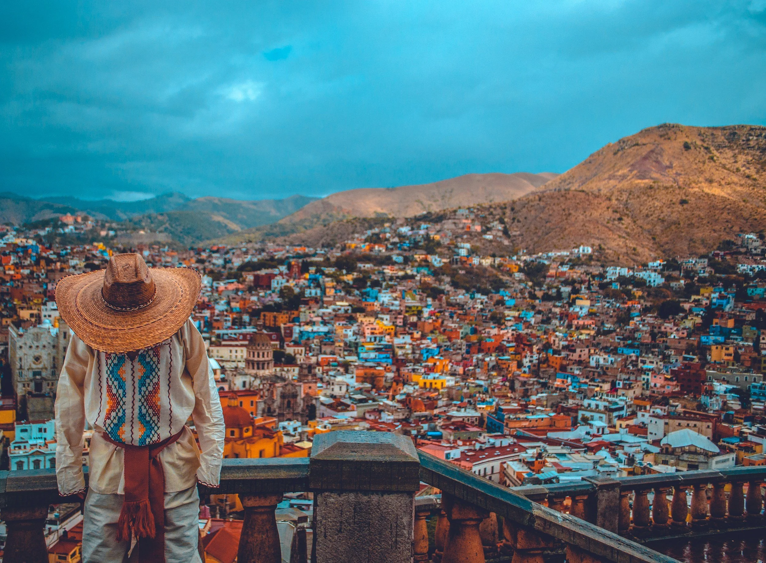 Person in traditional Mexican attire with a large straw hat, overlooking a colorful cityscape with hills in the background.