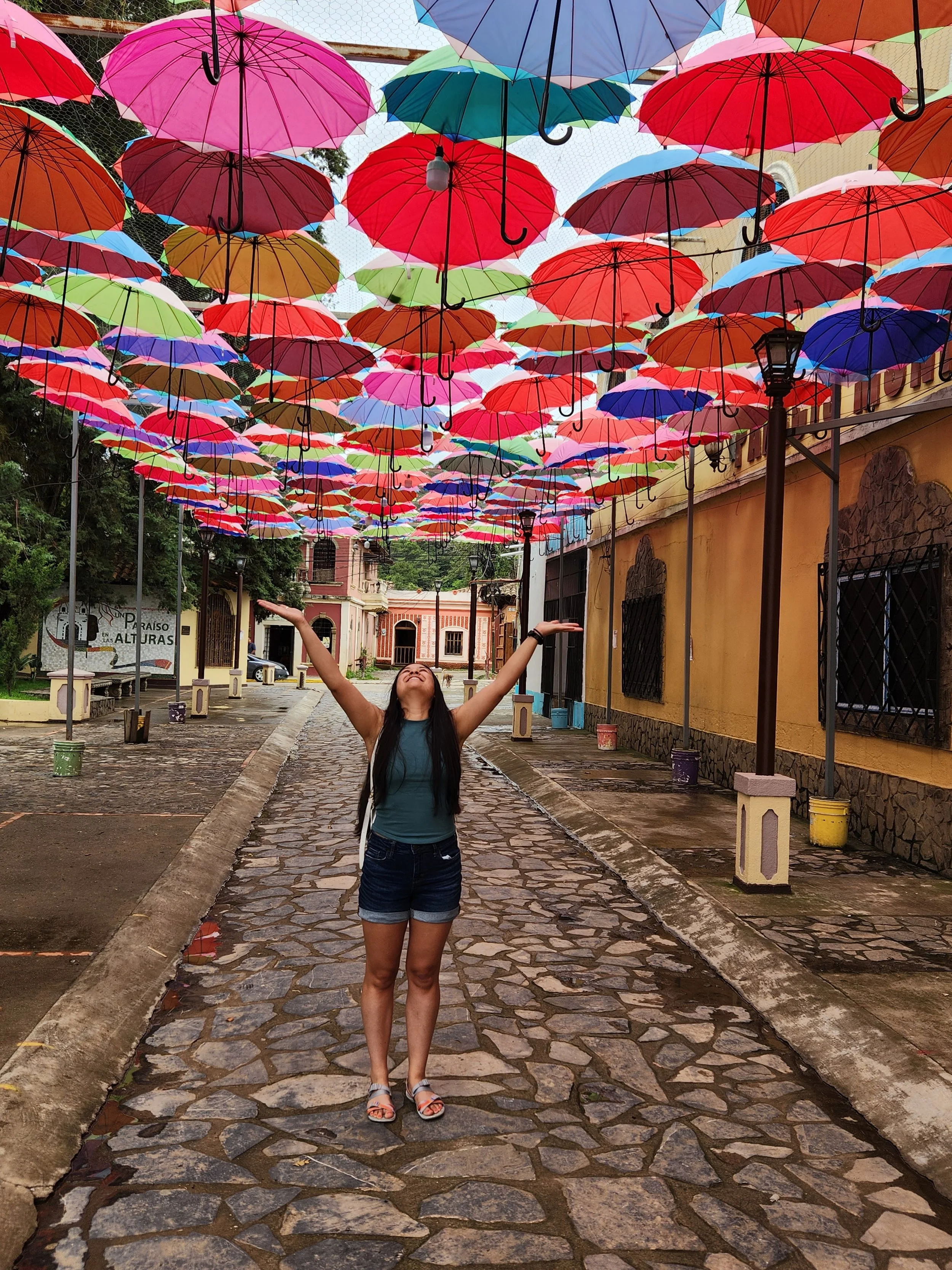 A woman standing on a cobblestone street with her arms raised under a colorful canopy of umbrellas hanging above her.