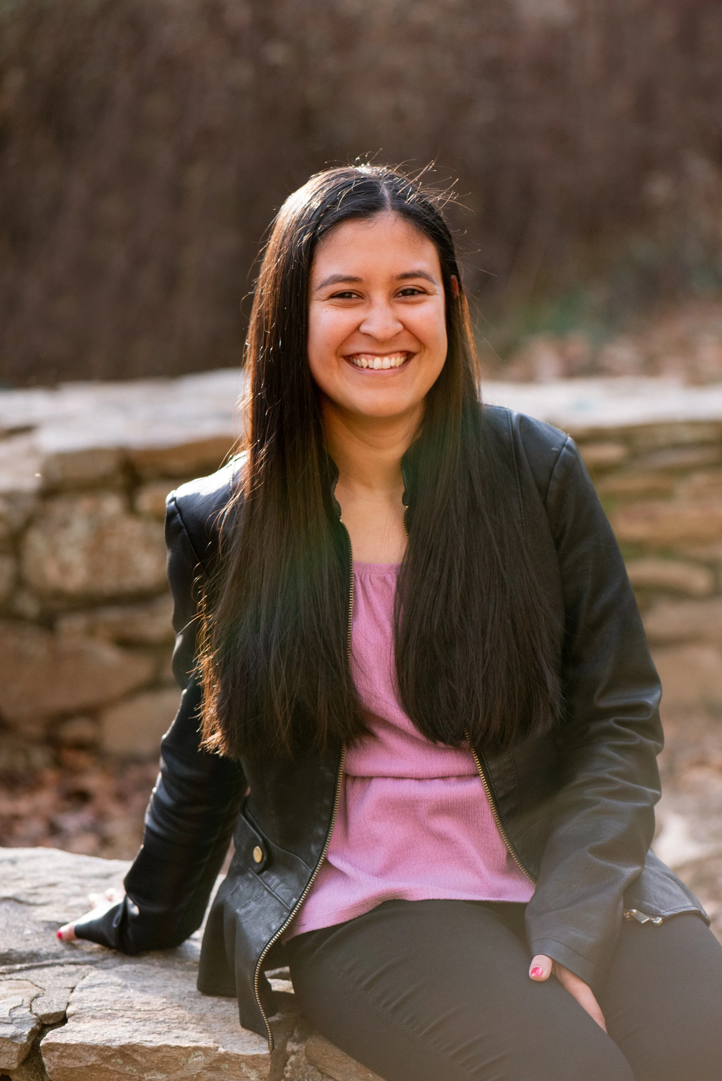 A woman with long dark hair is sitting outdoors on a stone ledge, smiling at the camera. She is wearing a black leather jacket over a pink top.