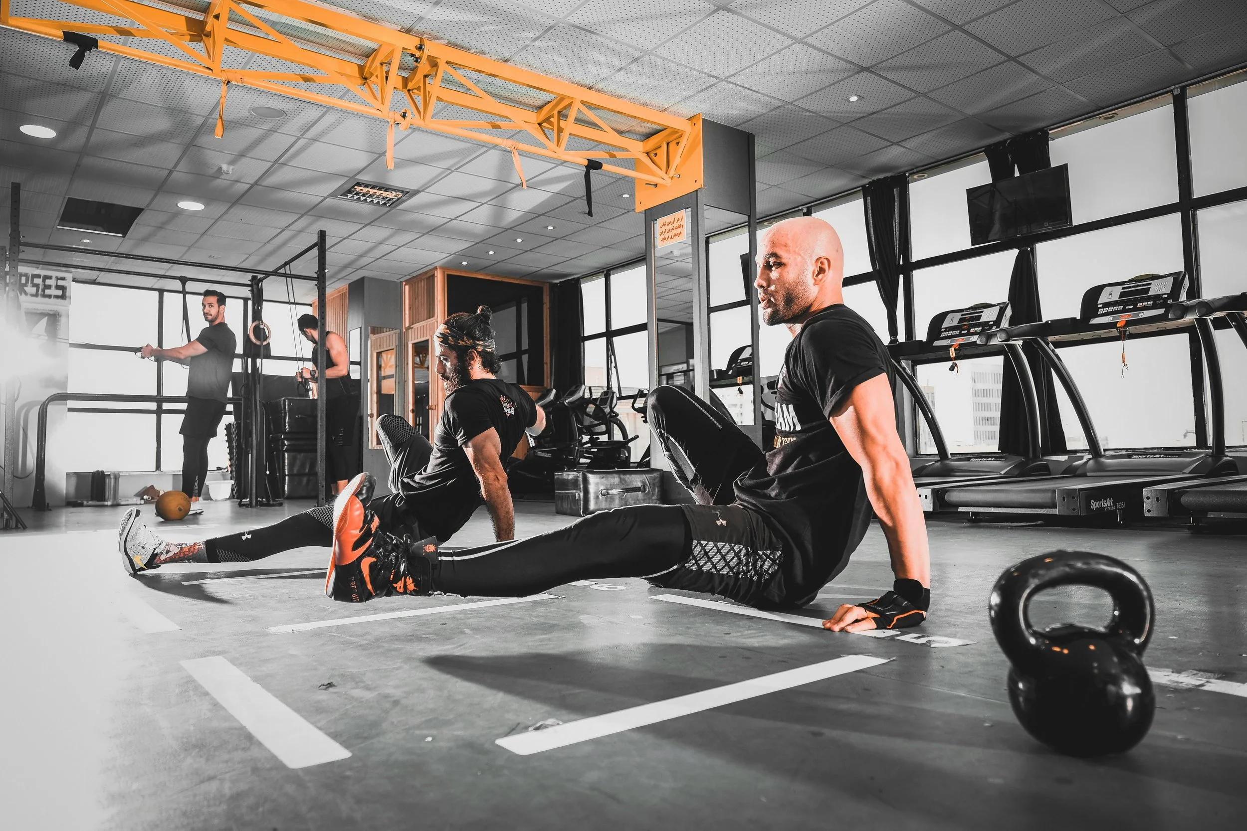 Two men performing floor exercises with one using his hands to lift his upper body off the ground and the other sitting with legs extended. The gym features treadmills, a kettlebell on the floor, and a bright interior with large windows and a yellow ceiling beam.
