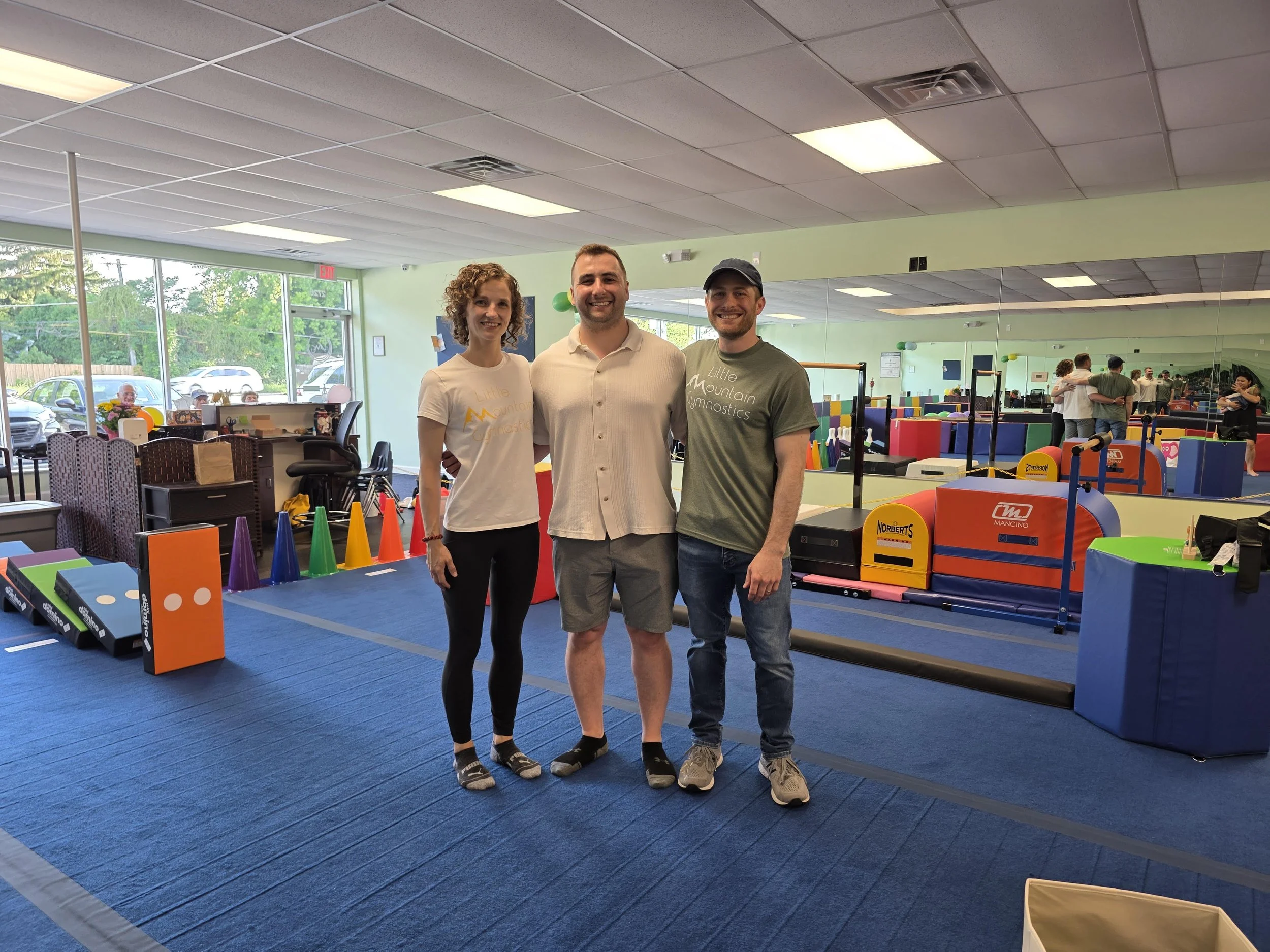 Three people standing together inside a gymnastics training facility, smiling at the camera. The background features various colorful gymnastics equipment and a large mirror reflecting other people training.