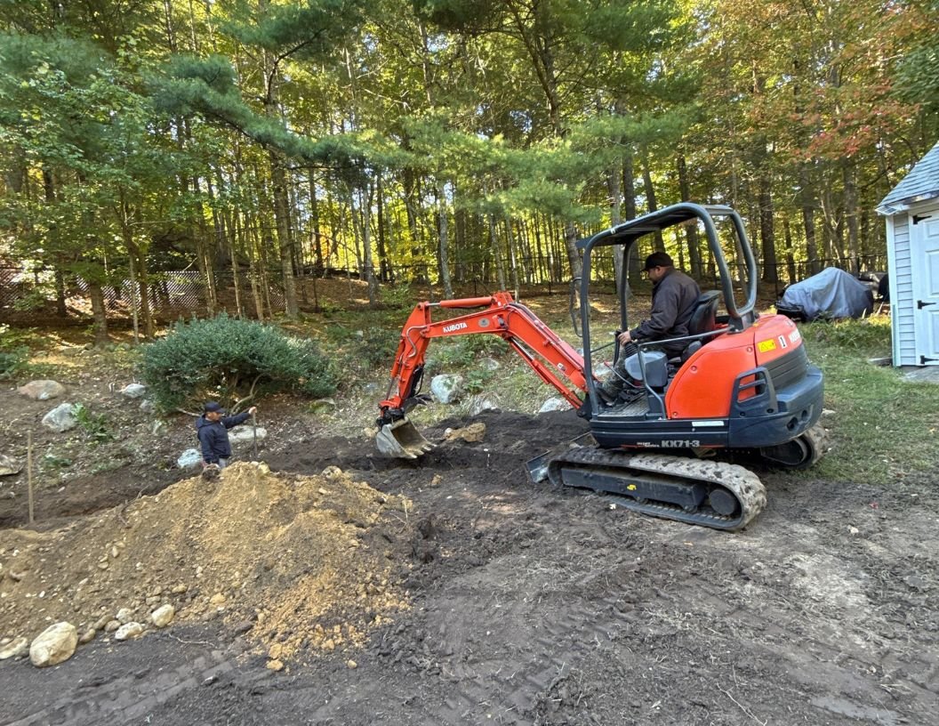 Two people working outside, one operating a small orange excavator and the other standing nearby on uneven dirt ground surrounded by trees.