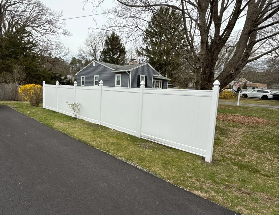 A white vinyl privacy fence along a suburban street with a blue house in the background and trees without leaves.