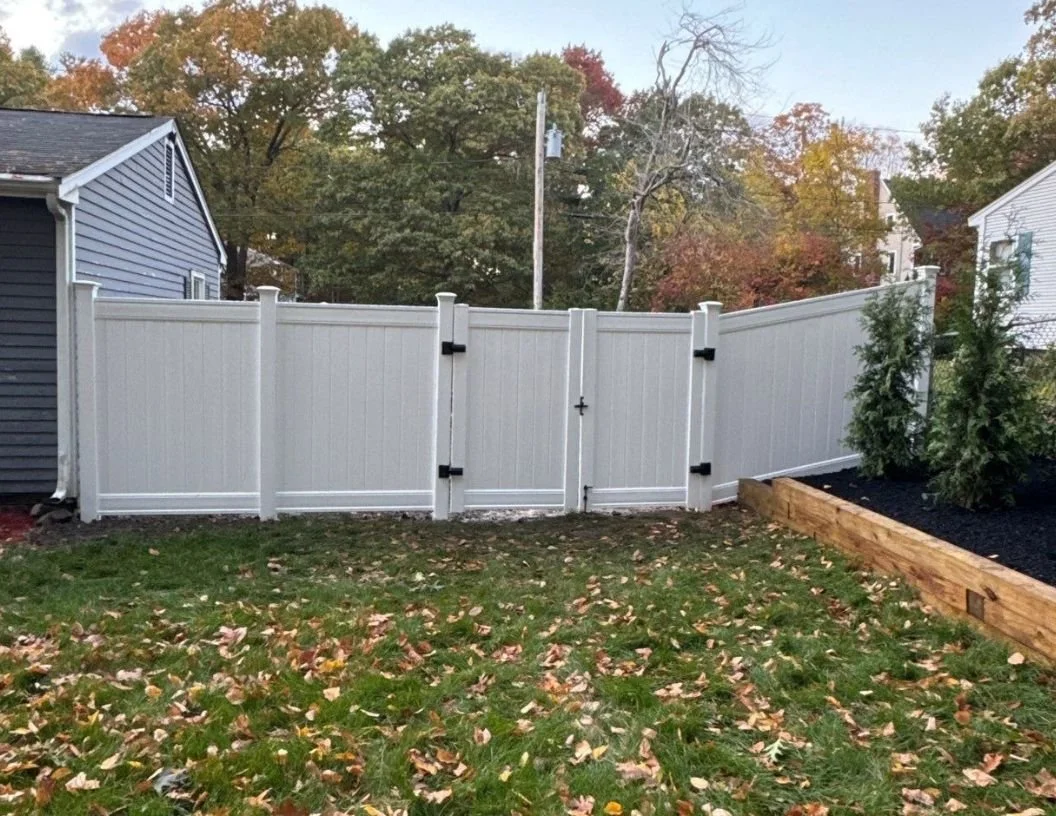 View of a white vinyl privacy fence with a gate, next to a garden bed with evergreen shrubs and a black mulch ground covering, in a backyard with fall leaves on the grass and trees with autumn foliage in the background.