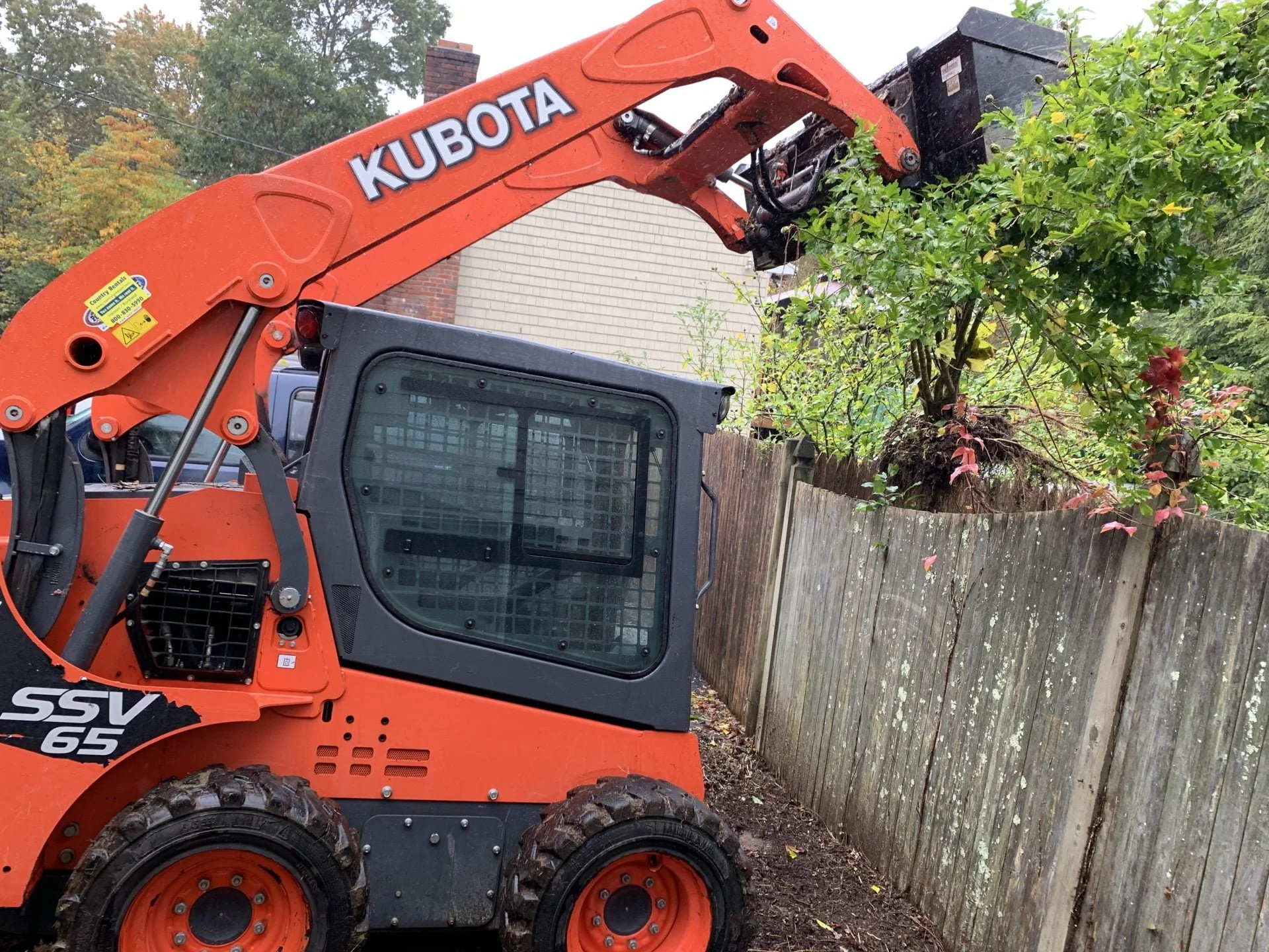 A Kubota skid-steer loader lifting a large shrub or small tree over a wooden fence in a residential yard.