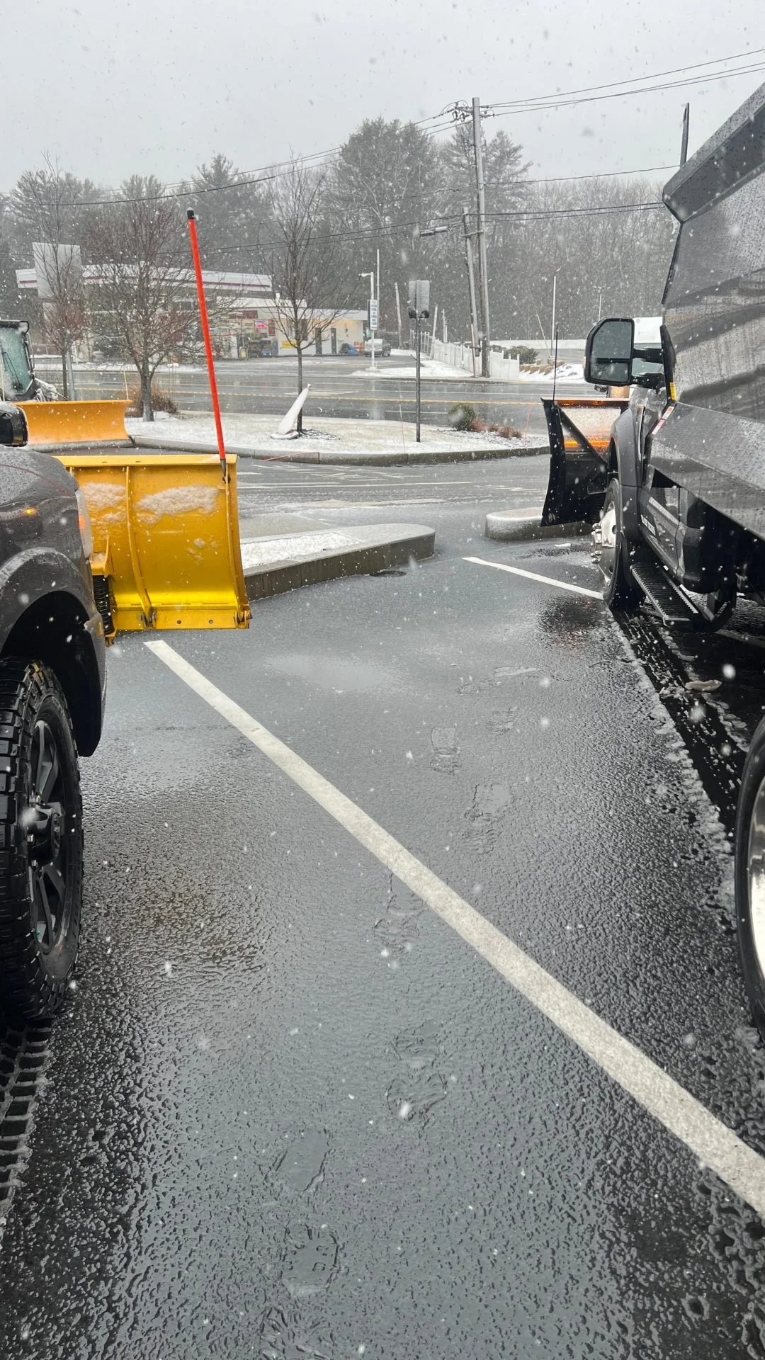 Snow falling in a parking lot with a snow plow attached to a truck on the right and another vehicle with a yellow snow blade on the left, wet asphalt, tire tracks, and commercial buildings in the background.