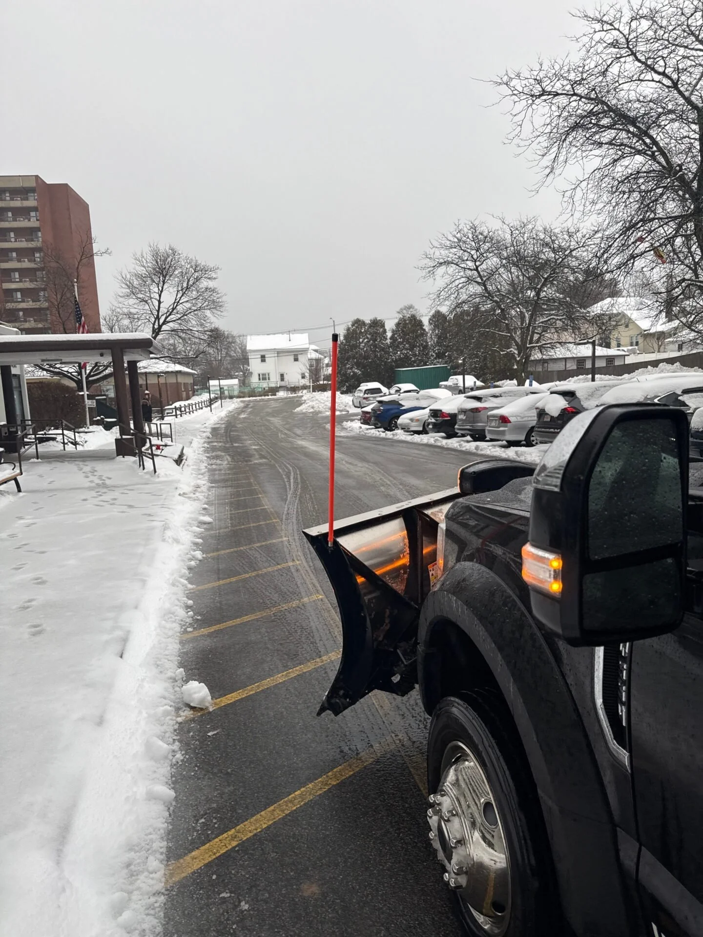 A snow plow truck clearing snow from a parking lot during winter with snow-covered cars and leafless trees in the background.