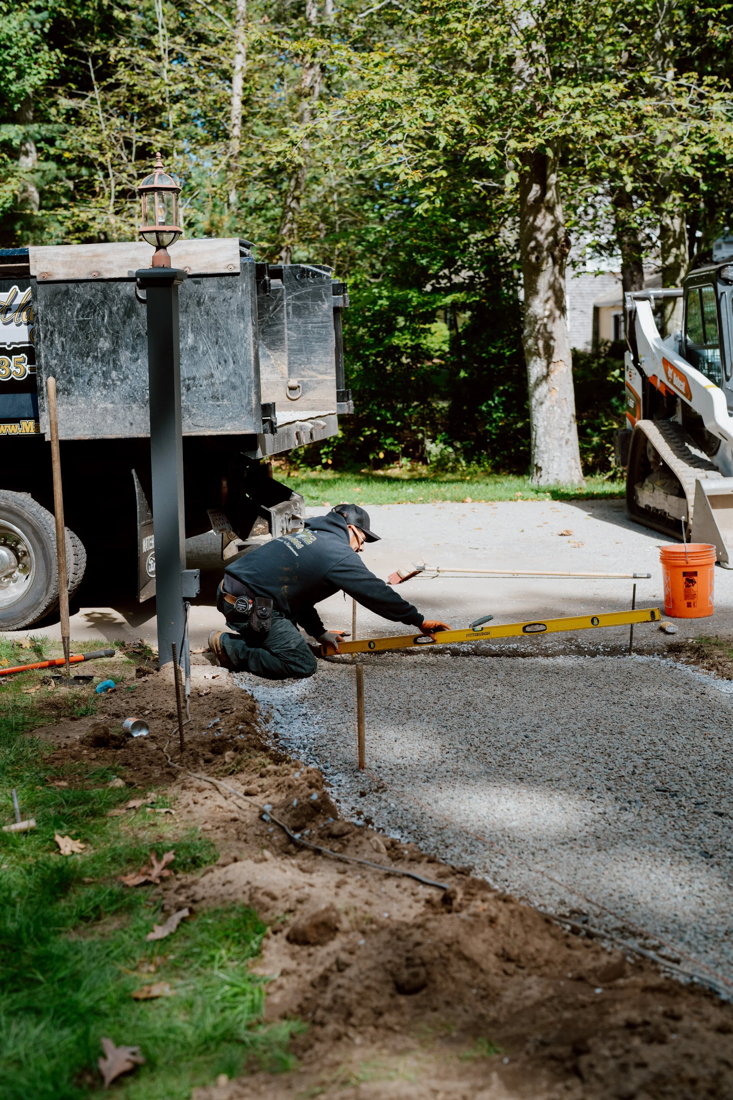 A worker leveling freshly poured concrete on a sidewalk during a construction project in a residential area with trees in the background.