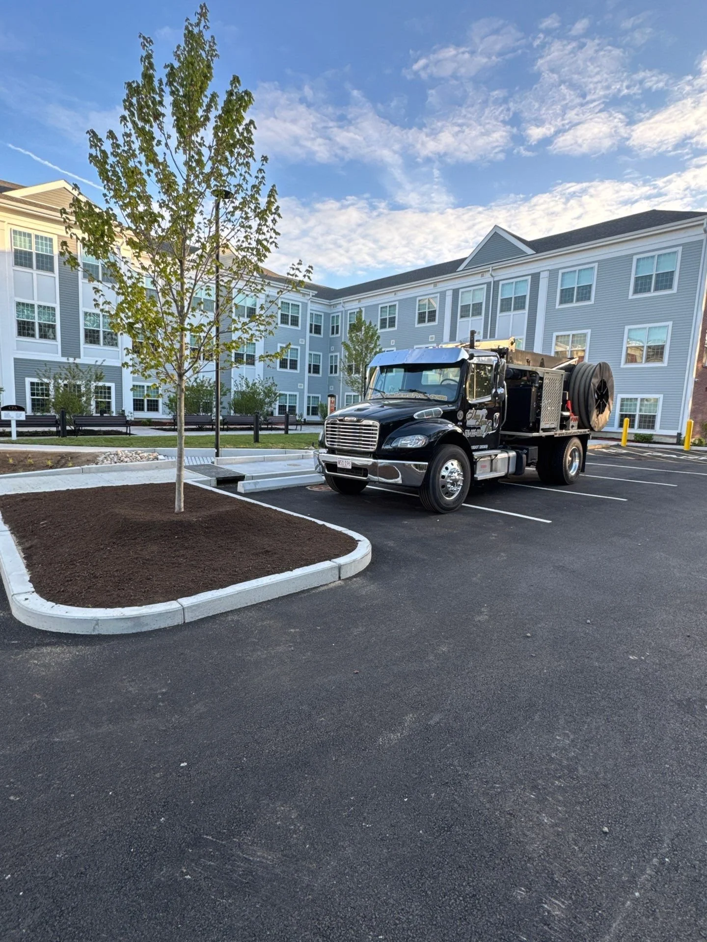 A black utility truck parked in a parking lot next to a newly planted tree and apartment buildings in the background under a blue sky with clouds.