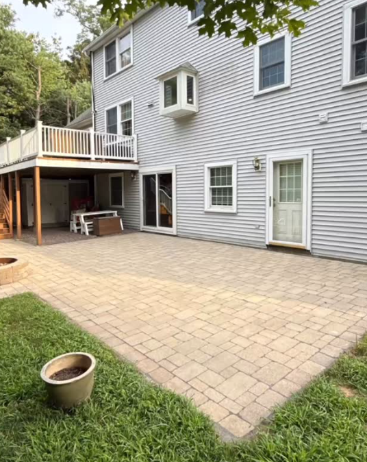 Backyard patio area with a newly paved brick surface, a small grassy patch with a flowerpot, a white door, and windows on a gray house with a deck above.