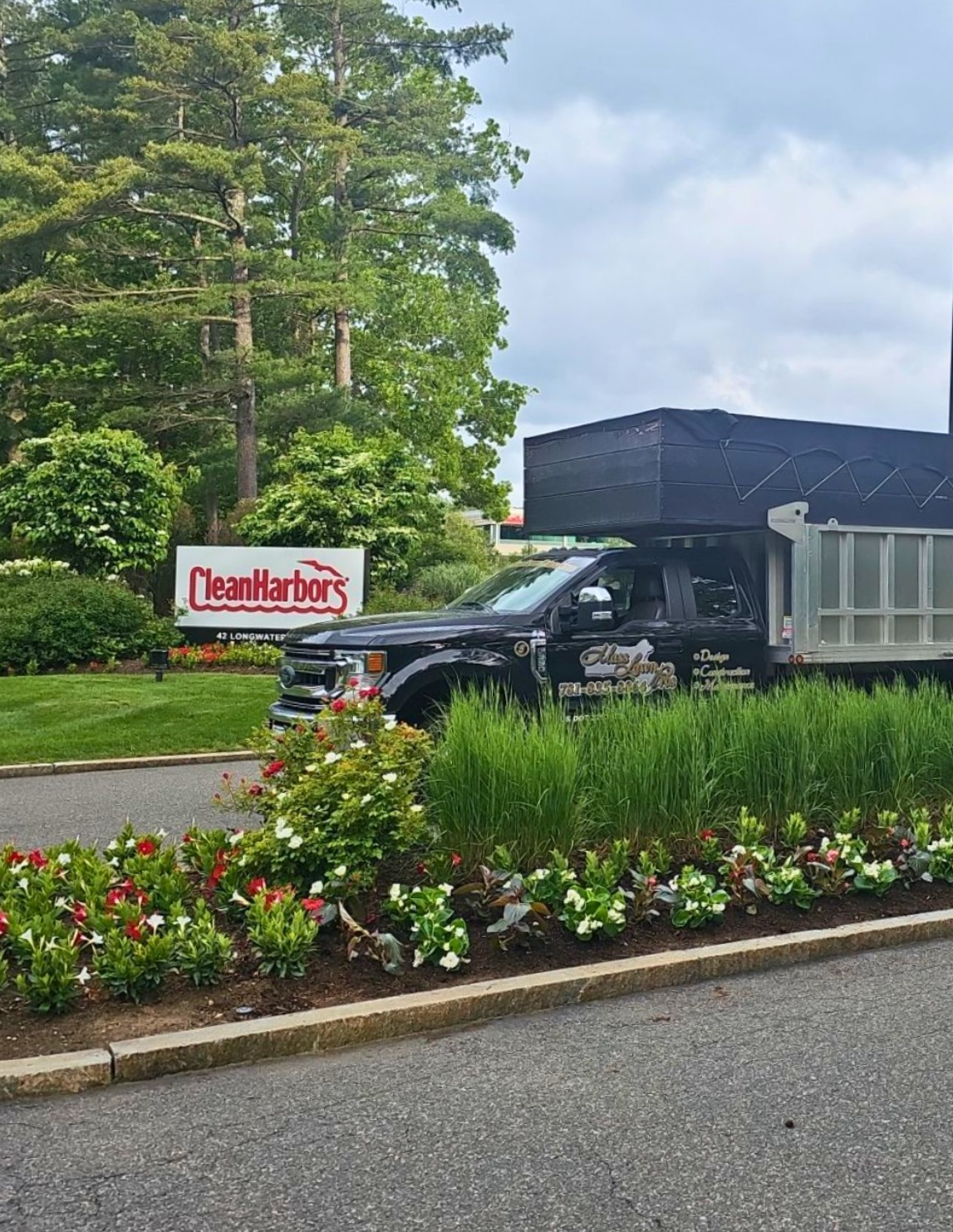 A black work truck parked on a driveway in front of a well-maintained flower bed with various flowers and green grass. In the background, there is a sign that reads 'Clean Harbors' and a row of tall trees.