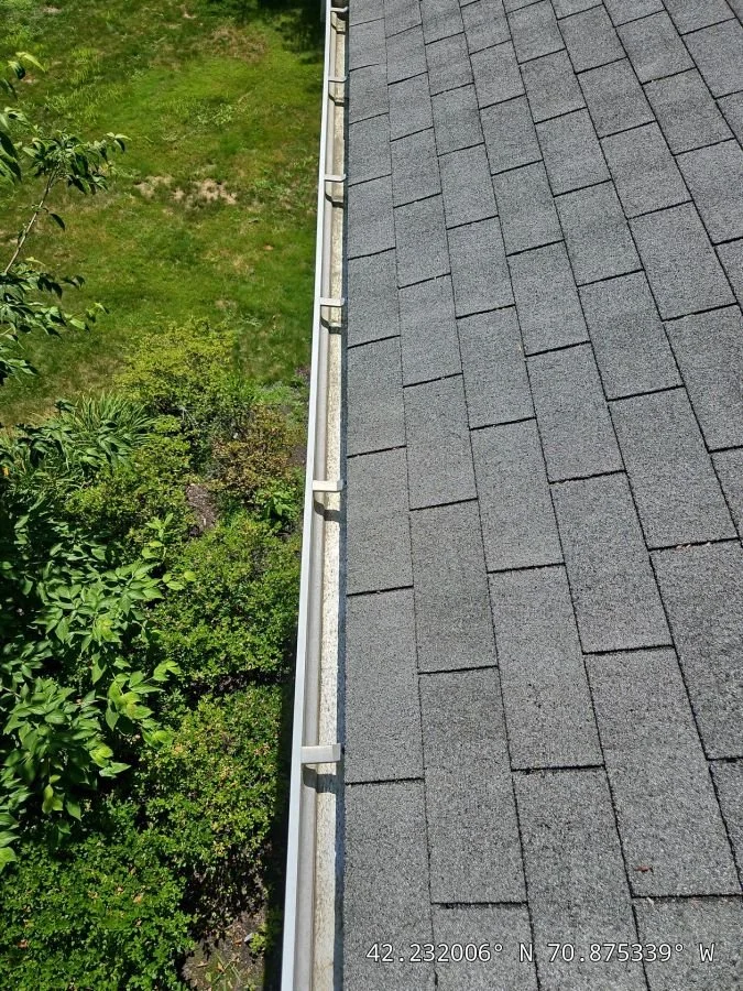 View of a roof with gray asphalt shingles and a gutter system, next to a green yard with grass and bushes.
