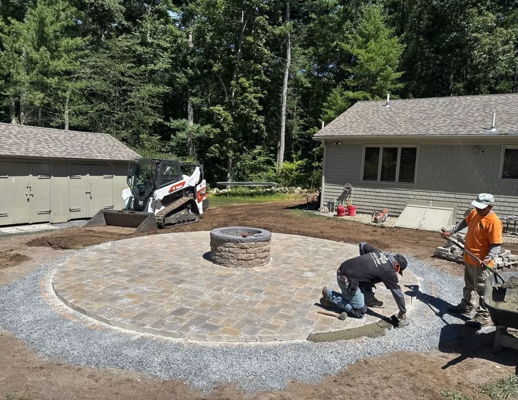 Workers laying pavers around a fire pit in a backyard, with a skid-steer loader nearby and a house in the background.