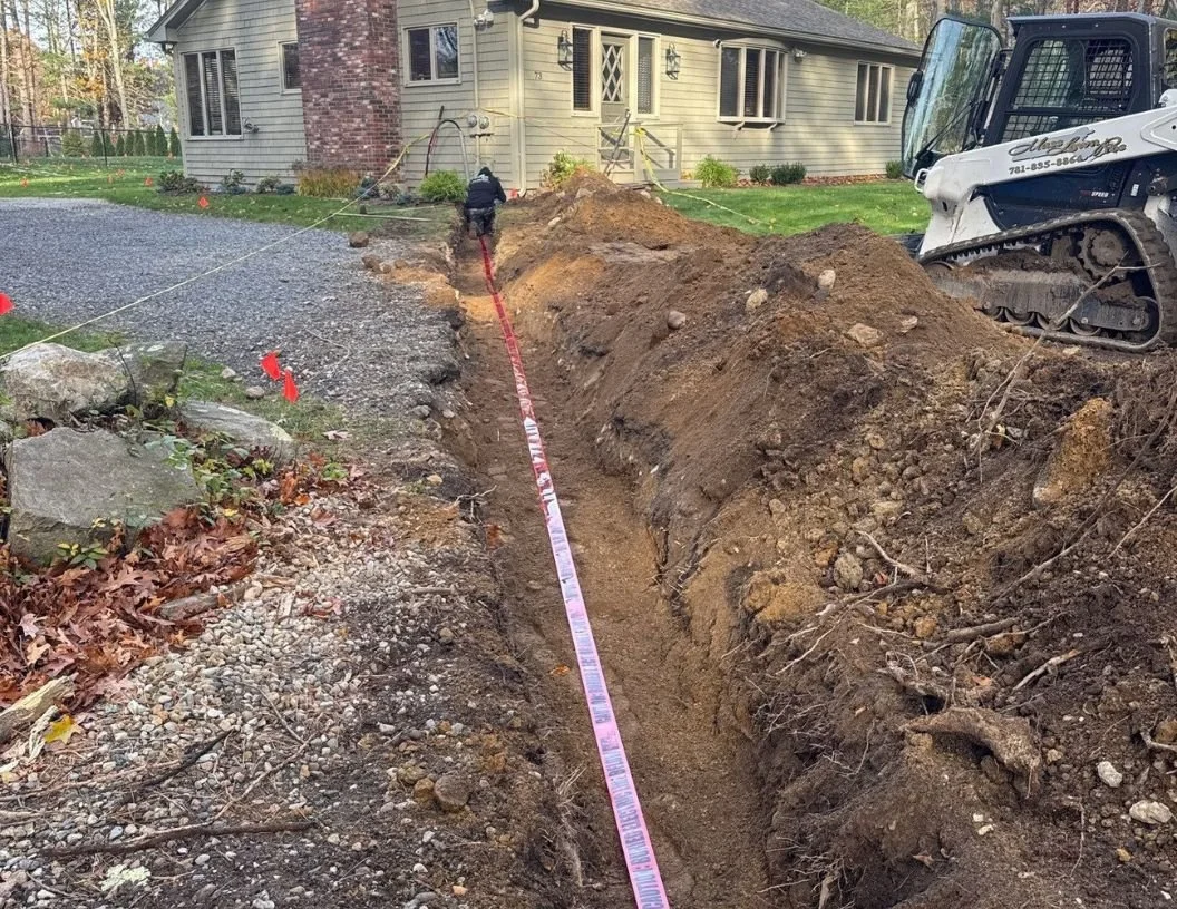 Construction workers digging a trench in front of a house for underground utility or irrigation work, with excavation equipment nearby.