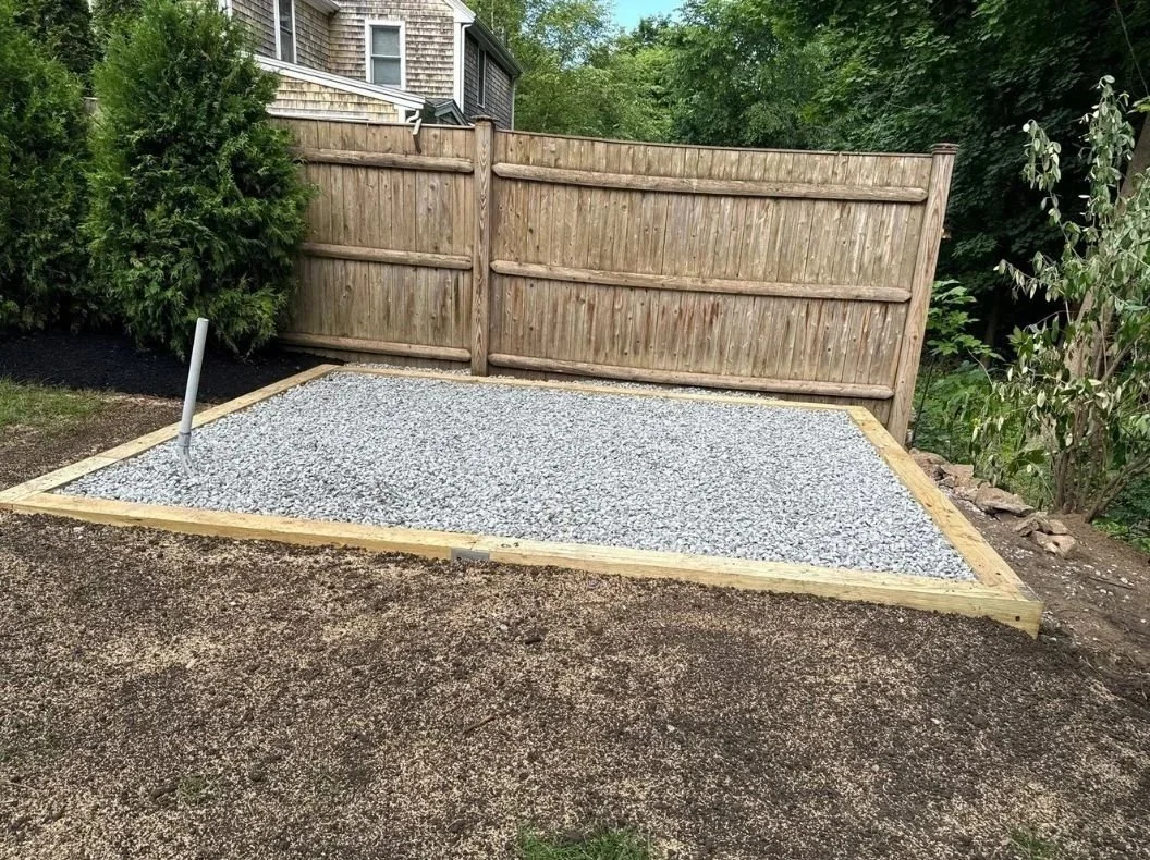 A backyard area with a gravel section bordered by wood framing, enclosed by a wooden fence, with trees and shrubs in the background.
