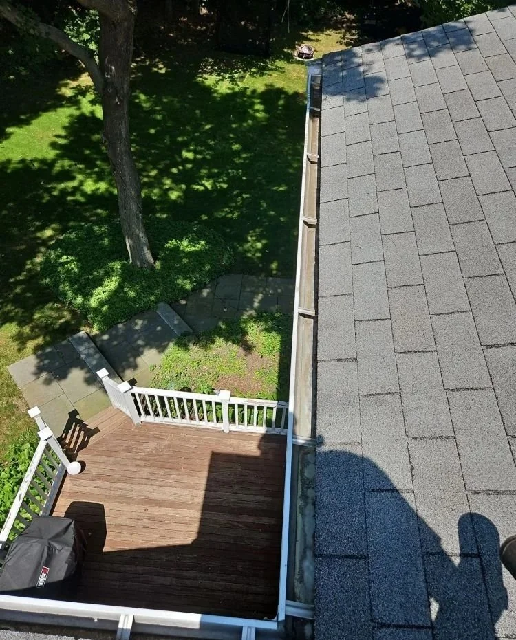 View from above of a backyard deck with white railing, a gray grill, and stairs leading to a grassy yard with a large tree. Part of the house roof with gray shingles is visible on the right side.
