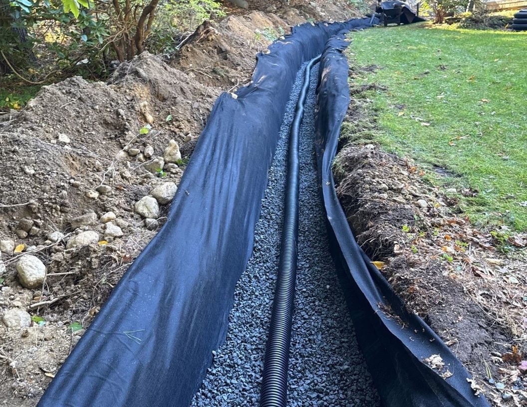 A drainage trench lined with black fabric and filled with gravel for underground piping, with a black corrugated pipe laid in the trench.