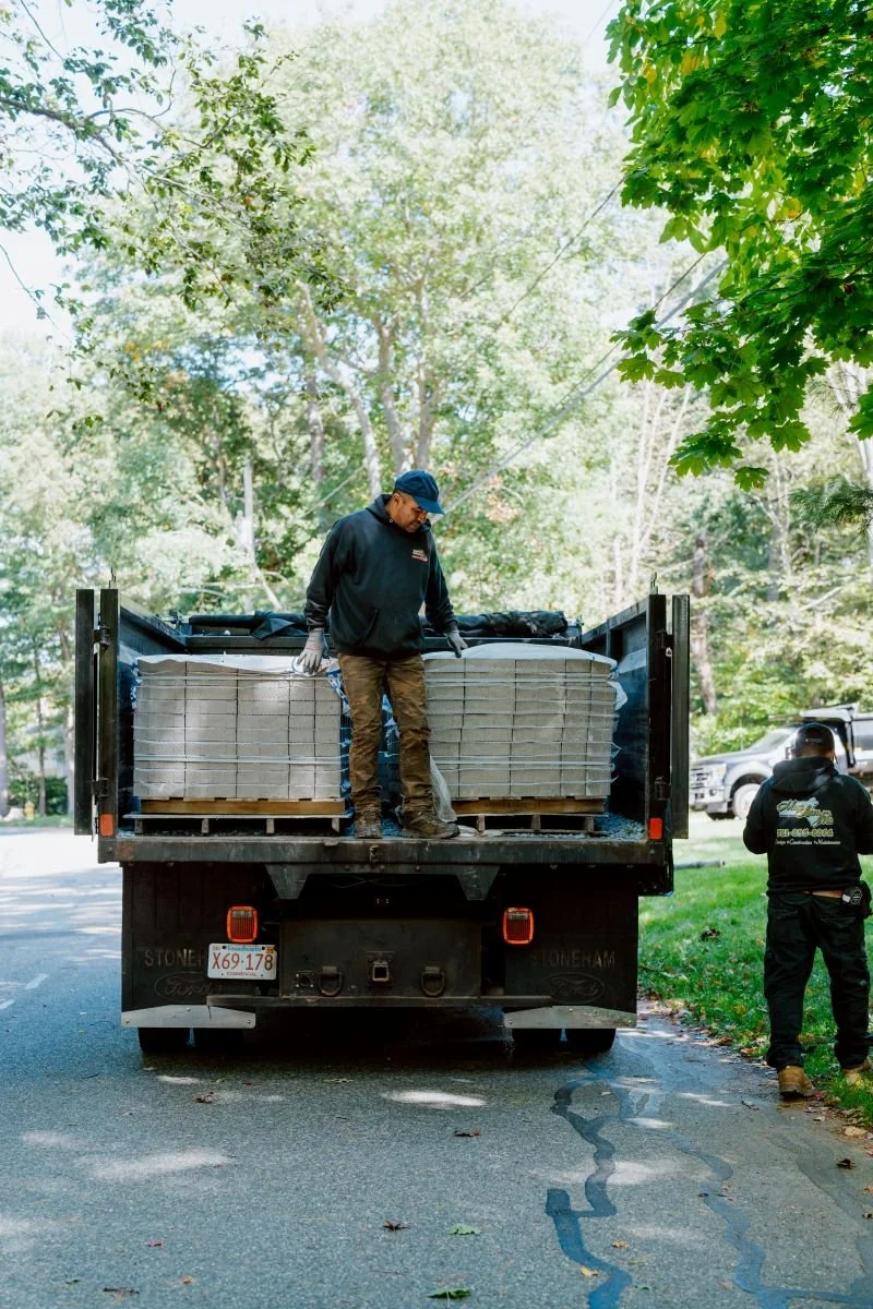 Two men unloading large construction materials from a black flatbed truck on a street lined with trees.