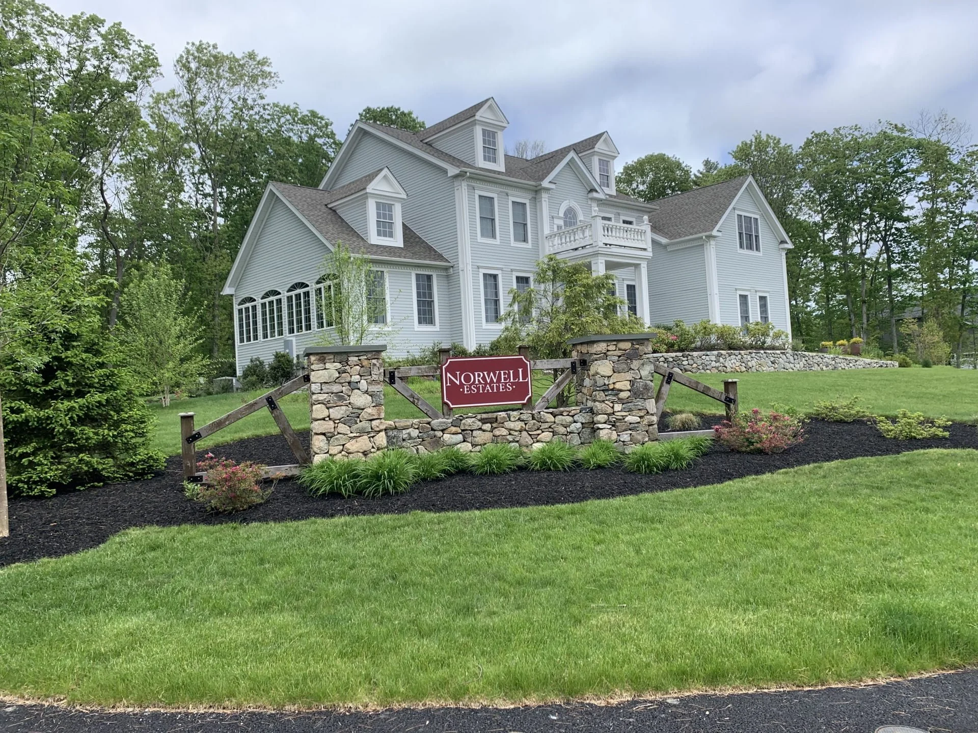 Large white house with multiple stories, gabled roofs, and a small balcony, surrounded by a well-maintained lawn and landscaping. A sign in front reads "Norwell Estates" supported by stone pillars and wooden fencing.