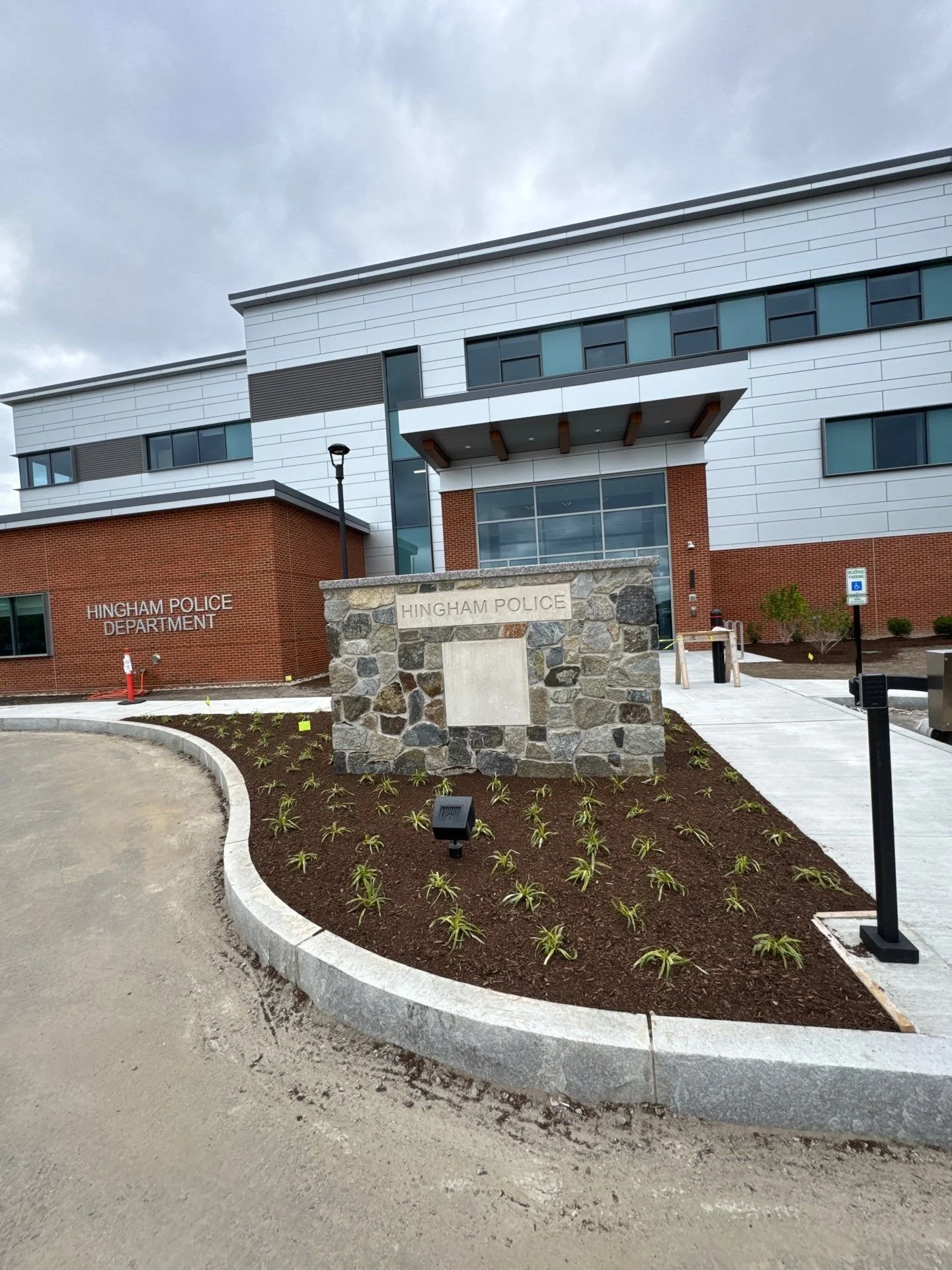Exterior view of the Hingham Police Department building with a stone sign in the front yard, landscaped with small plants, under a cloudy sky.