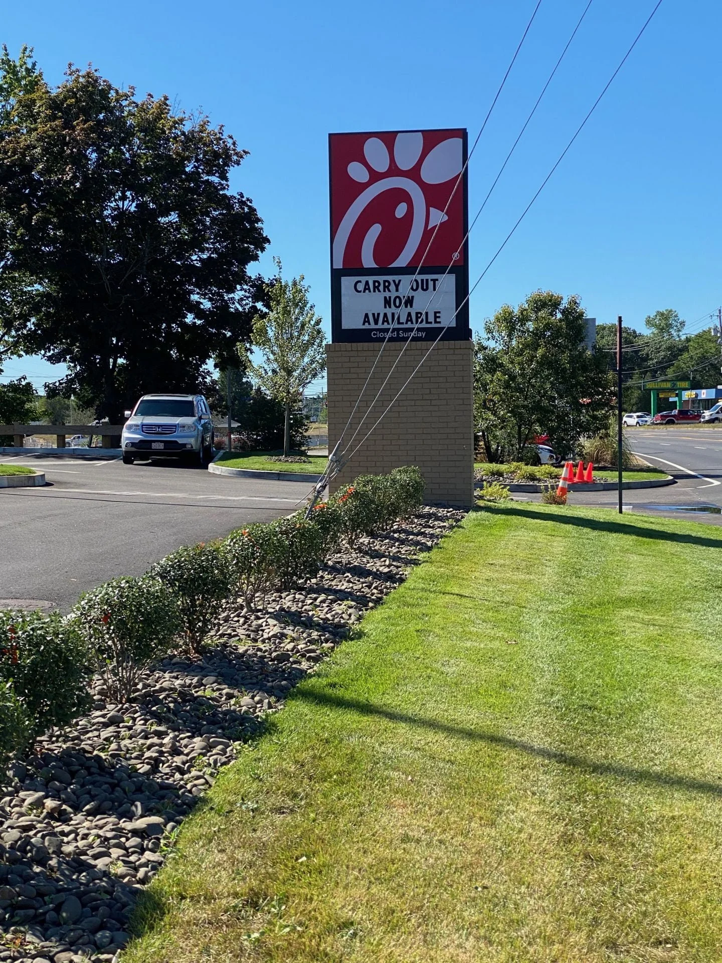 Chick-fil-A restaurant sign indicating carryout is now available and closed on Sundays, with a landscaped area and parked cars in the background.