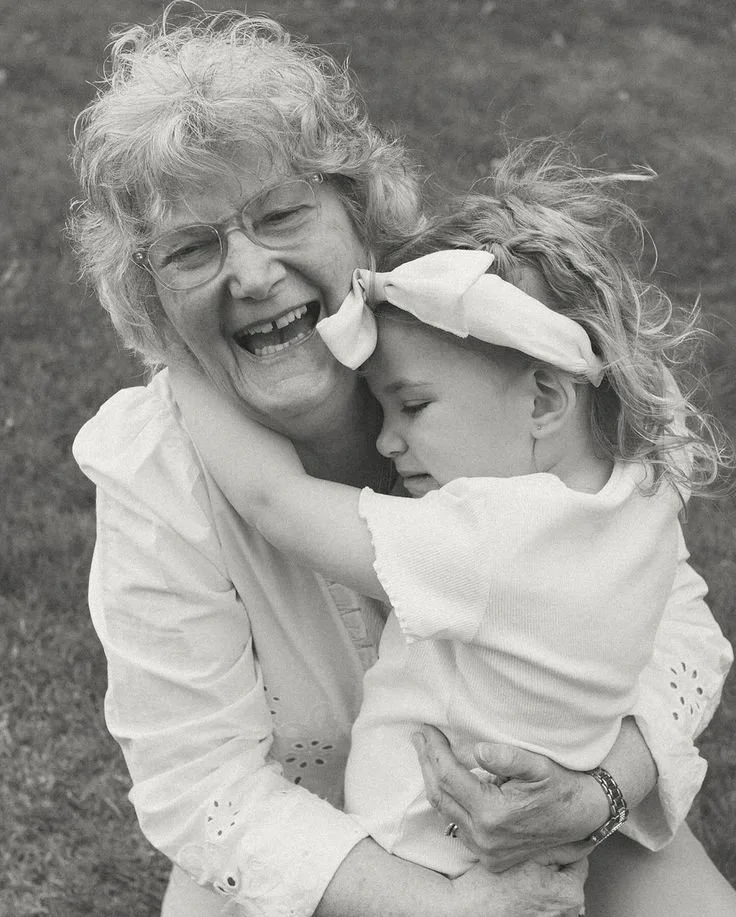 An elderly woman with glasses and curly hair warmly hugs and smiles with a young girl with a bow in her hair. The girl is hugging the woman tightly with her eyes closed. They are outdoors on grass.