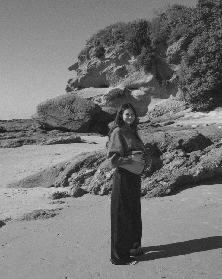A woman standing on a beach holding her pregnant belly, with large rocks and hills in the background, in black and white.