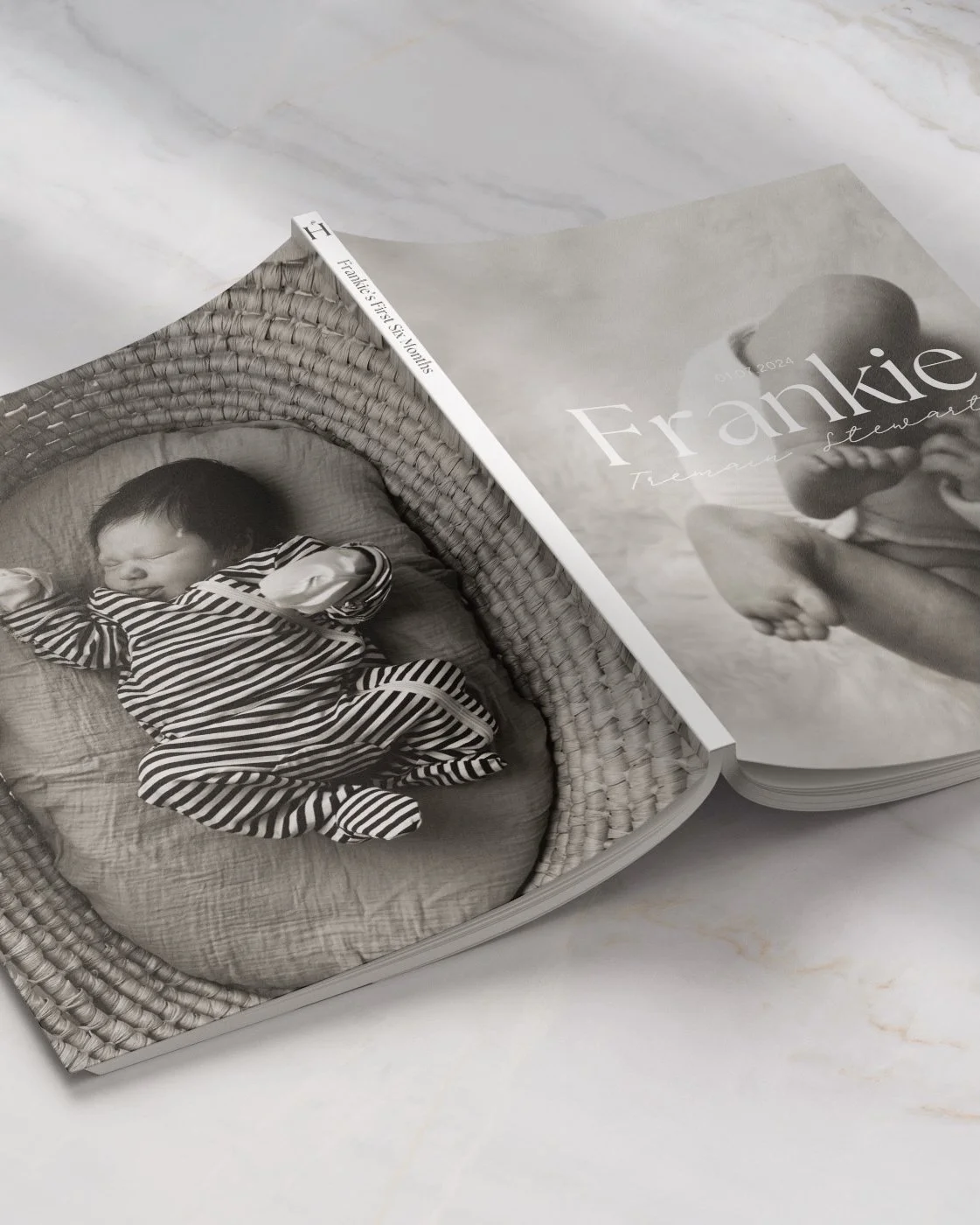A black and white photo of a sleeping baby in a striped outfit inside a woven basket, displayed on the pages of a photo book titled "Frankie".