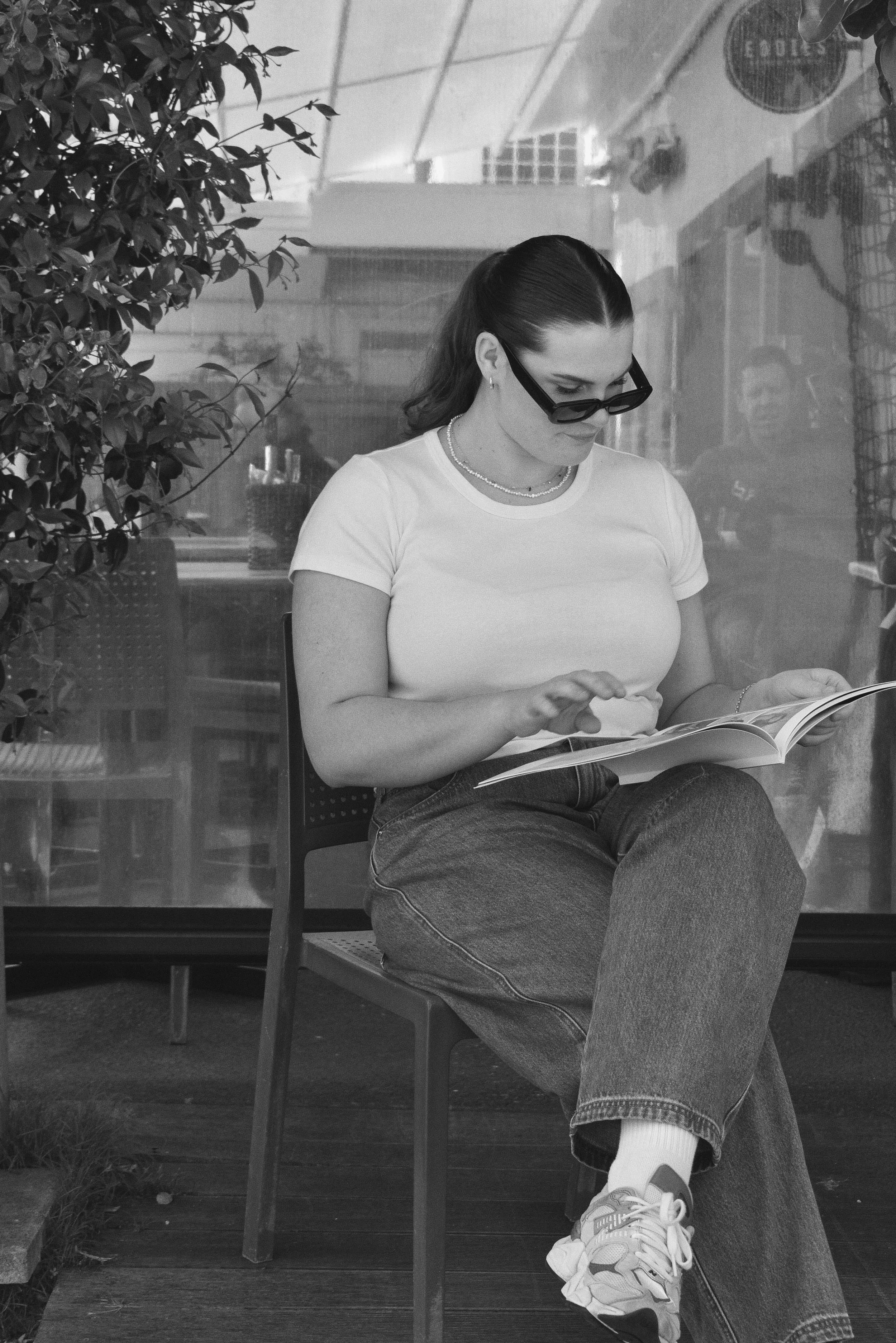 A woman sitting on a chair outdoors, reading a book, wearing sunglasses, a white t-shirt, jeans, and sneakers, with plants around her.