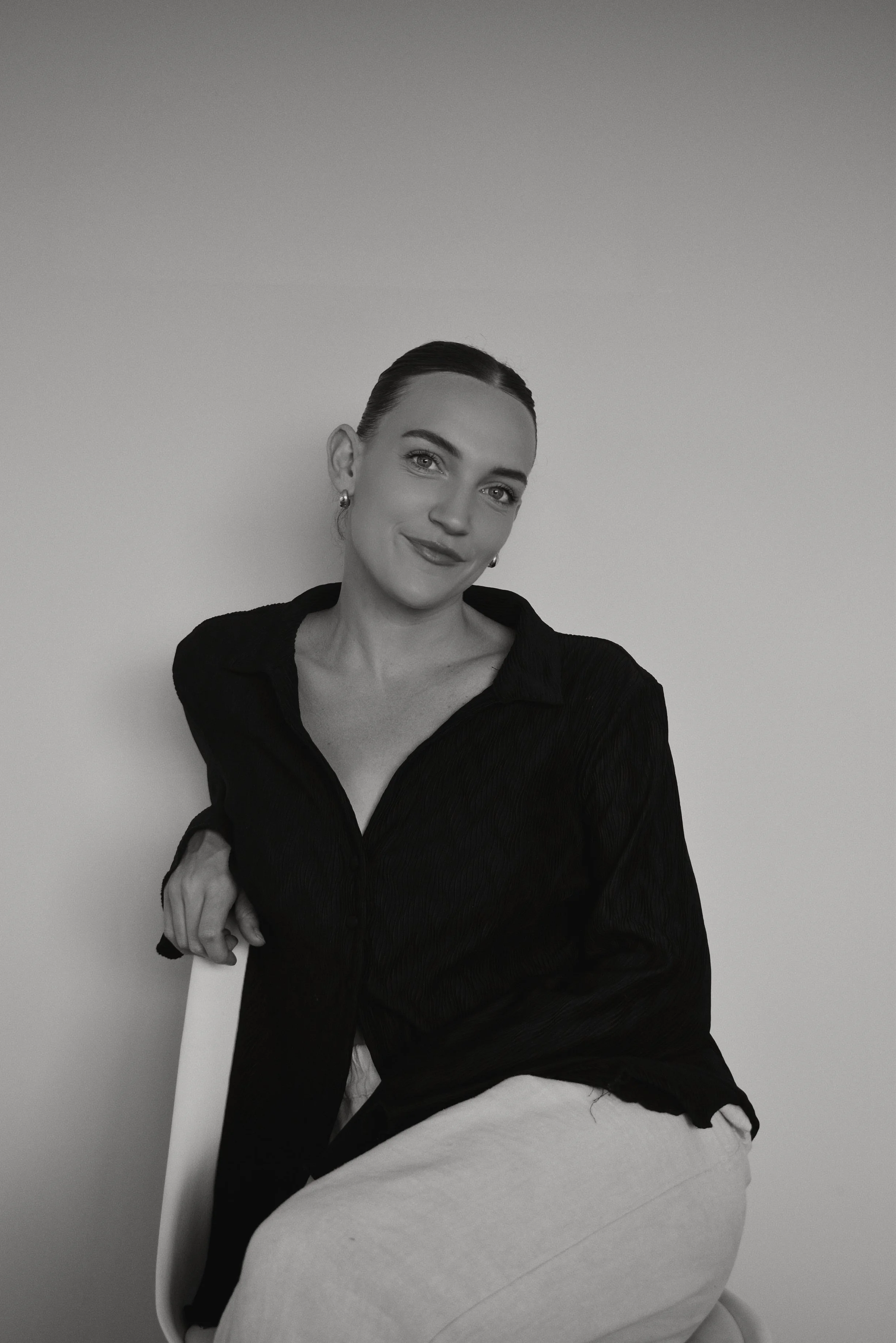 Black and white photo of a woman with dark hair pulled back, wearing a dark blouse and light-colored pants, sitting on a chair against a plain wall, smiling slightly.