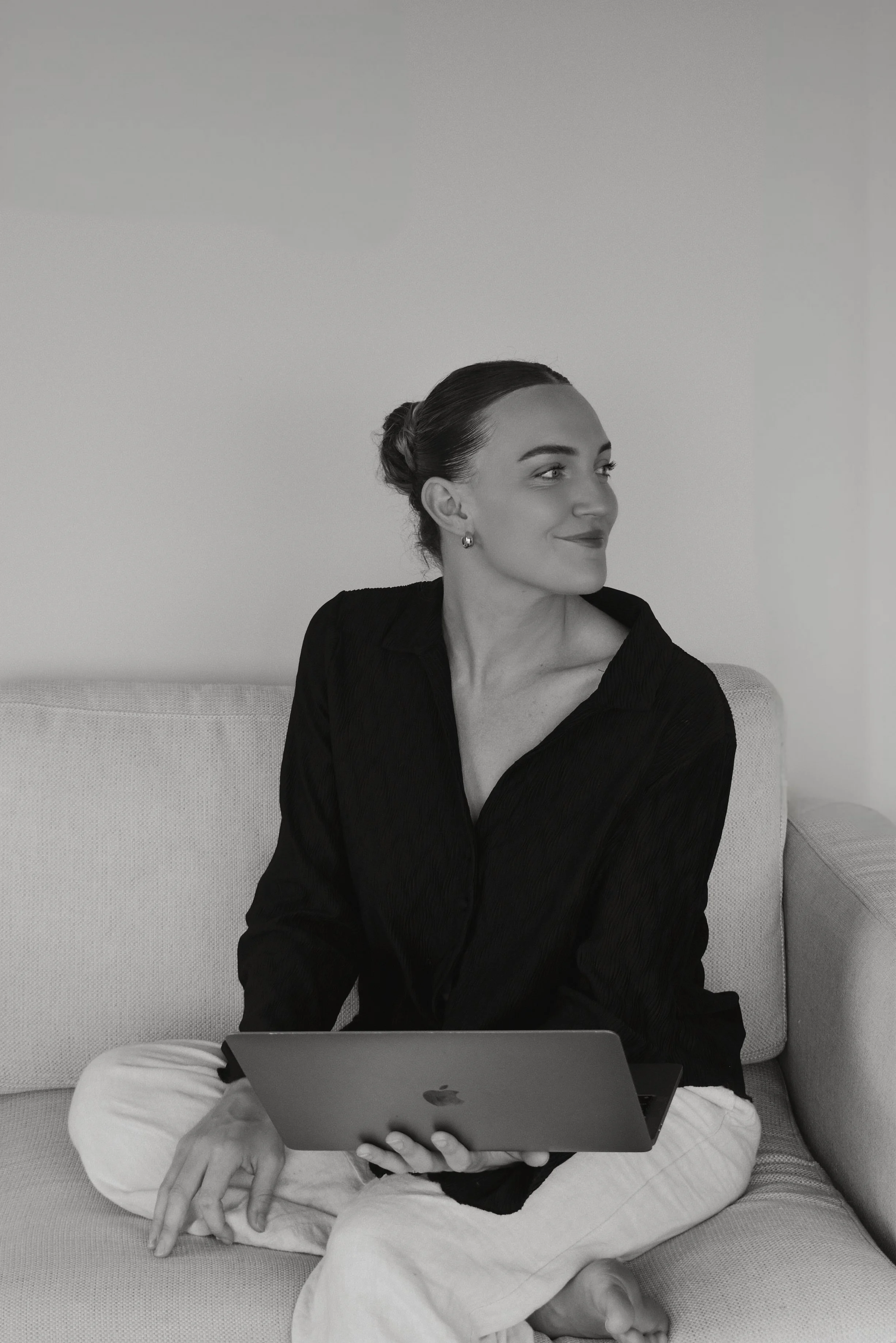 A woman sitting on a sofa holding a laptop, looking to the side, smiling, with her hair in a bun, wearing earrings, a black shirt, and light-colored pants, in a minimalist room.