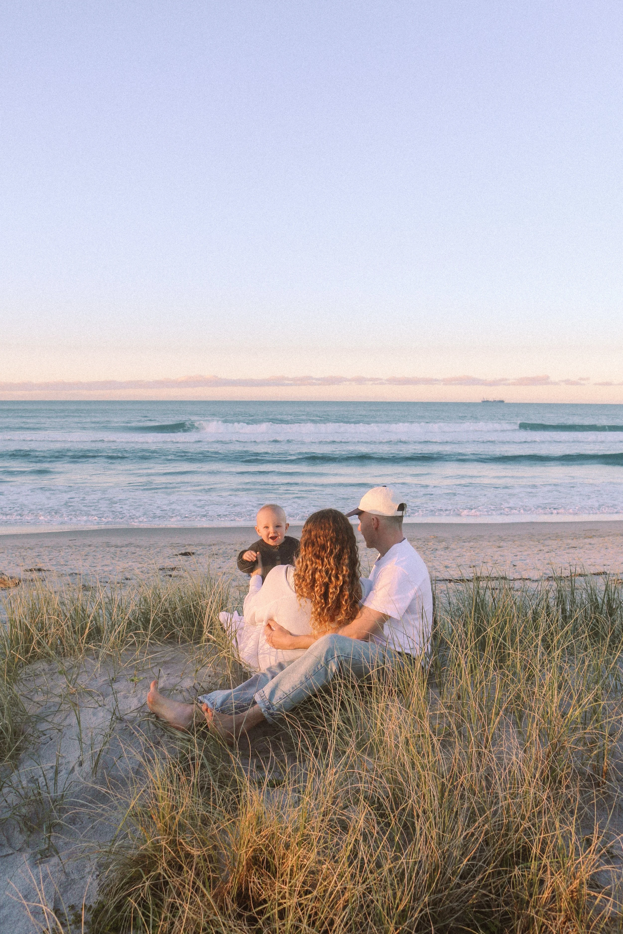 A family of three enjoying a beach sunset, sitting on sandy dunes among grass, with ocean waves and a ship in the background.
