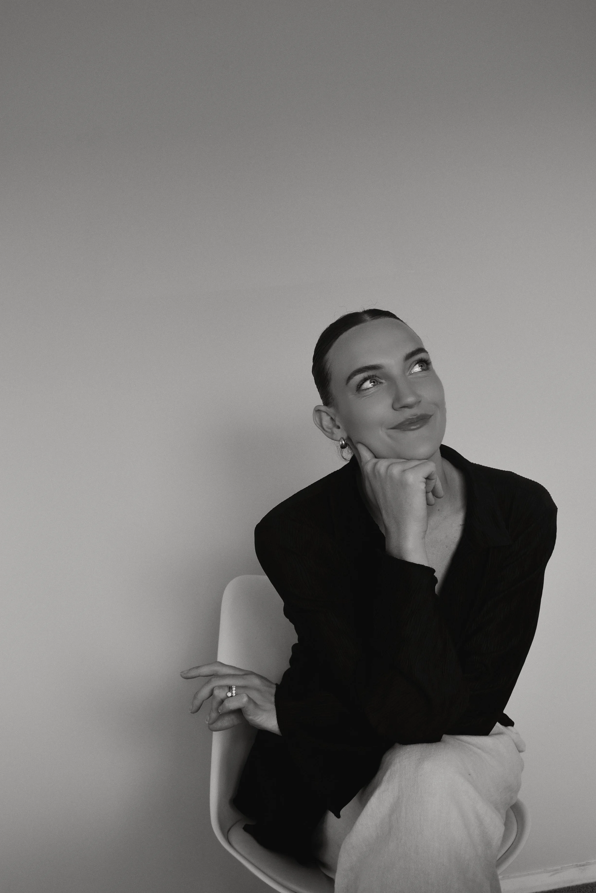Black and white photo of a woman sitting on a chair, resting her chin on her hand, with a thoughtful expression, looking upwards.