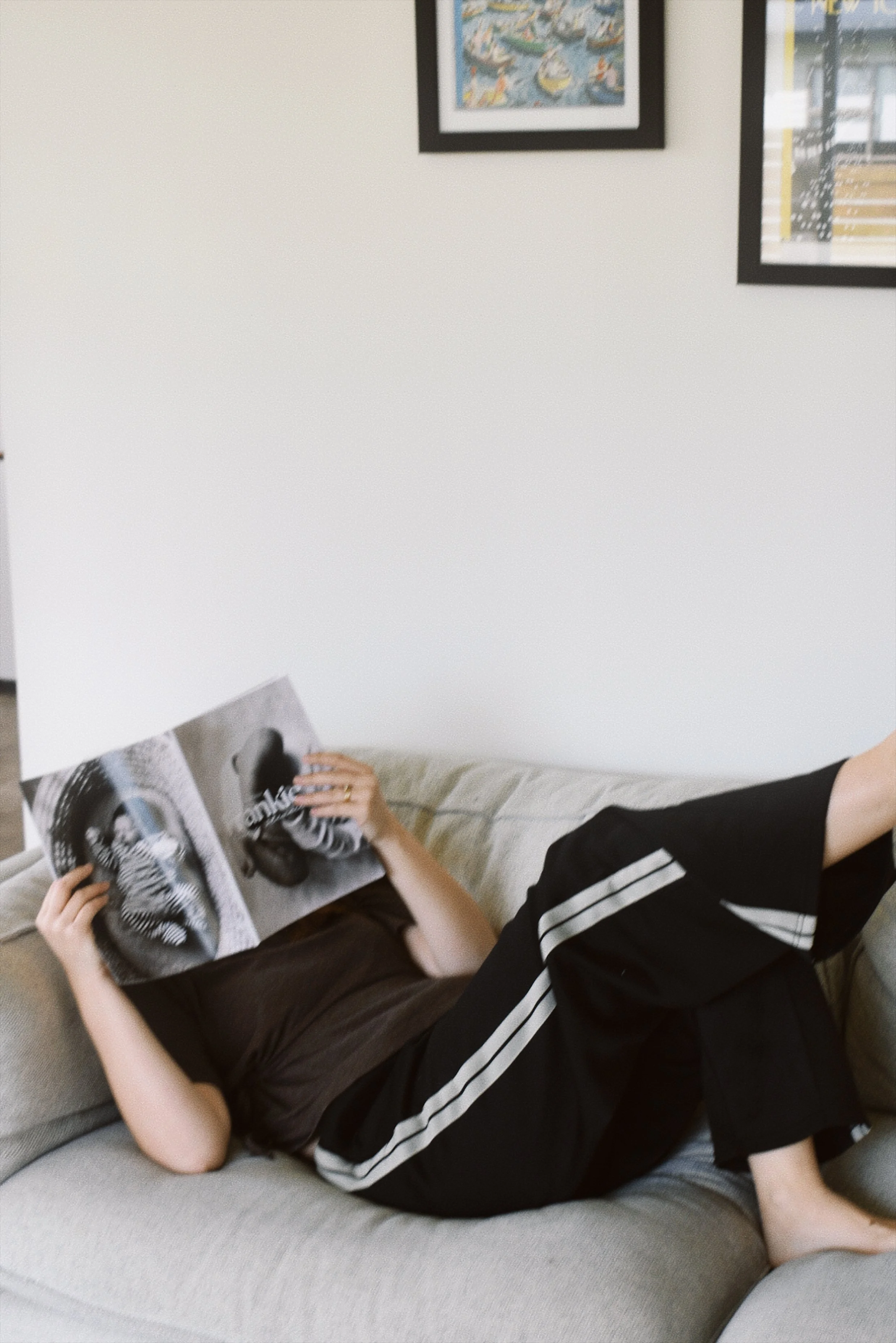 Person lying on a beige couch, reading a black and white photo magazine, wearing black pants with white stripes and a dark shirt, with artwork on the white wall behind.