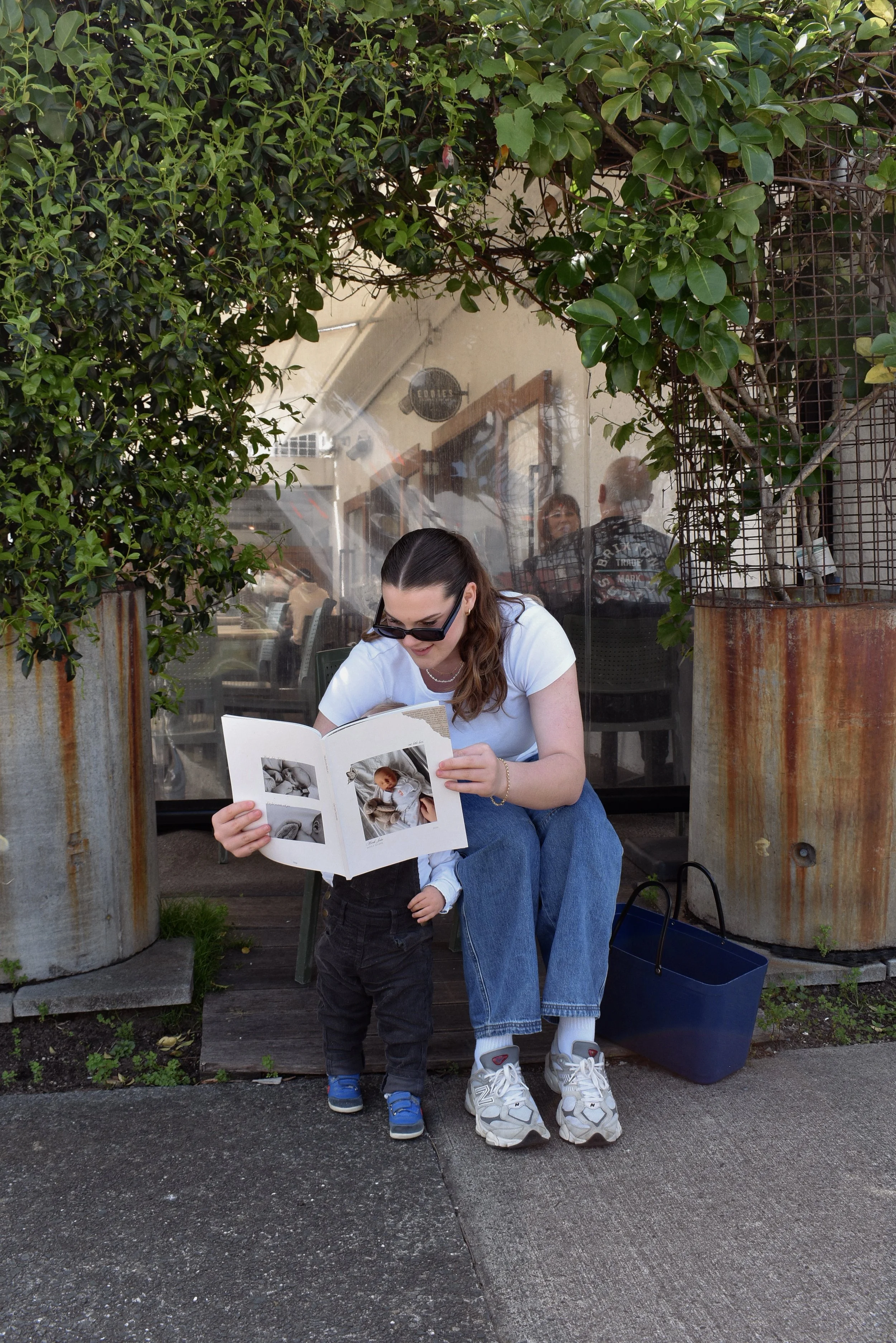 Young woman and small child looking at photographs, sitting outside a restaurant or café with greenery and rusted metal planters around them.