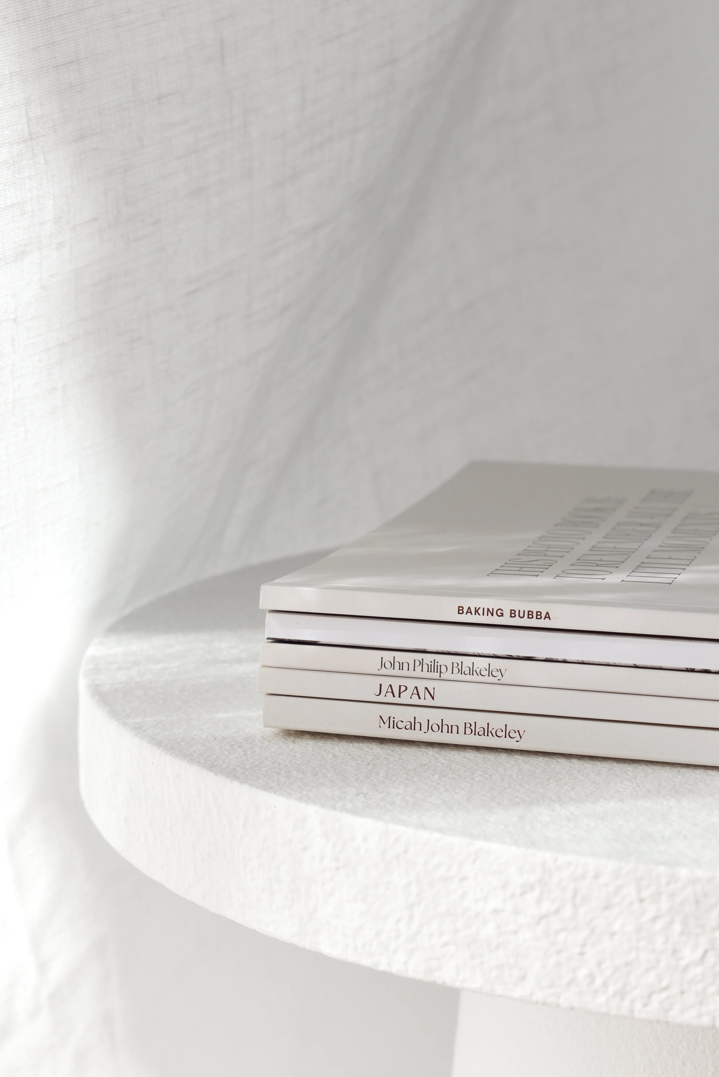 Stack of four white hardcover books on a rounded white shelf against a textured white wall.