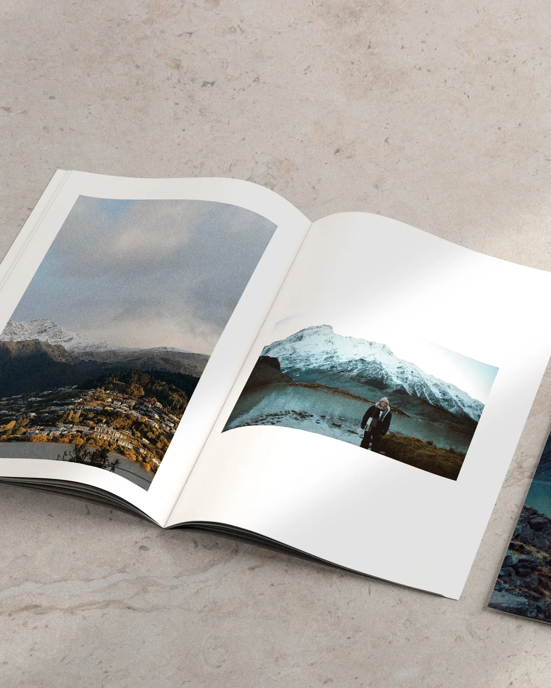 An open photo book showing images of mountains and a person standing near a lake with snow-capped peaks in the background, placed on a light-colored surface.
