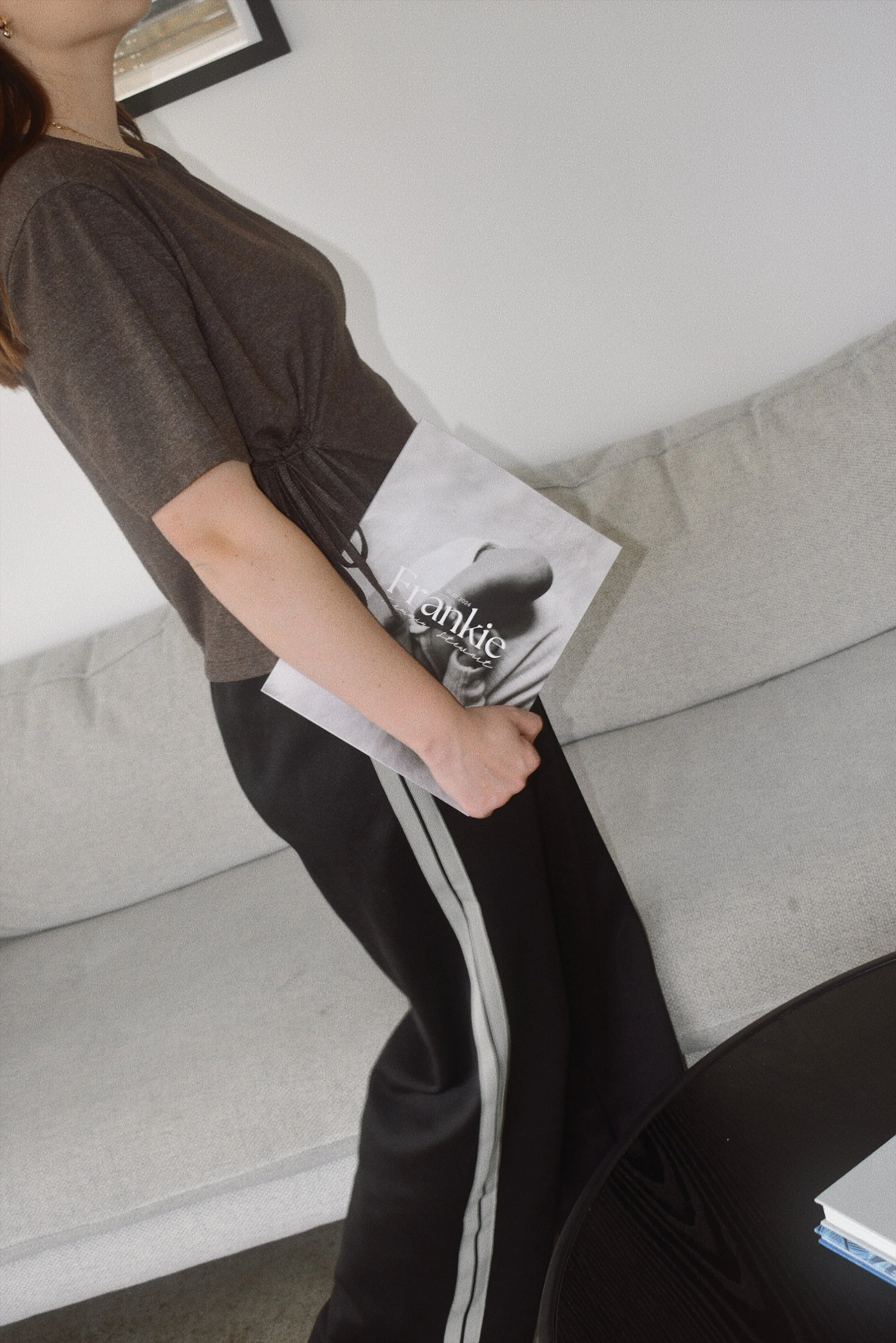 A woman is standing in a room, holding a magazine titled 'Frankie' featuring a black and white photo of a person on the cover. She is wearing a black t-shirt and black pants with white side stripes, next to a light-colored sofa and a small black tabl
