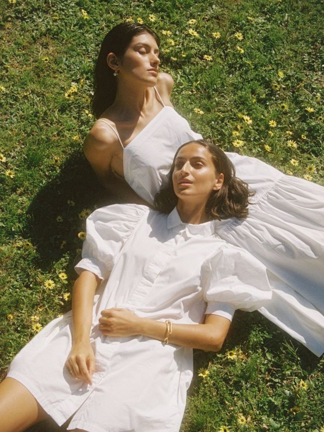 Two women lying on grass with yellow flowers, wearing white dresses, sunlit outdoors.