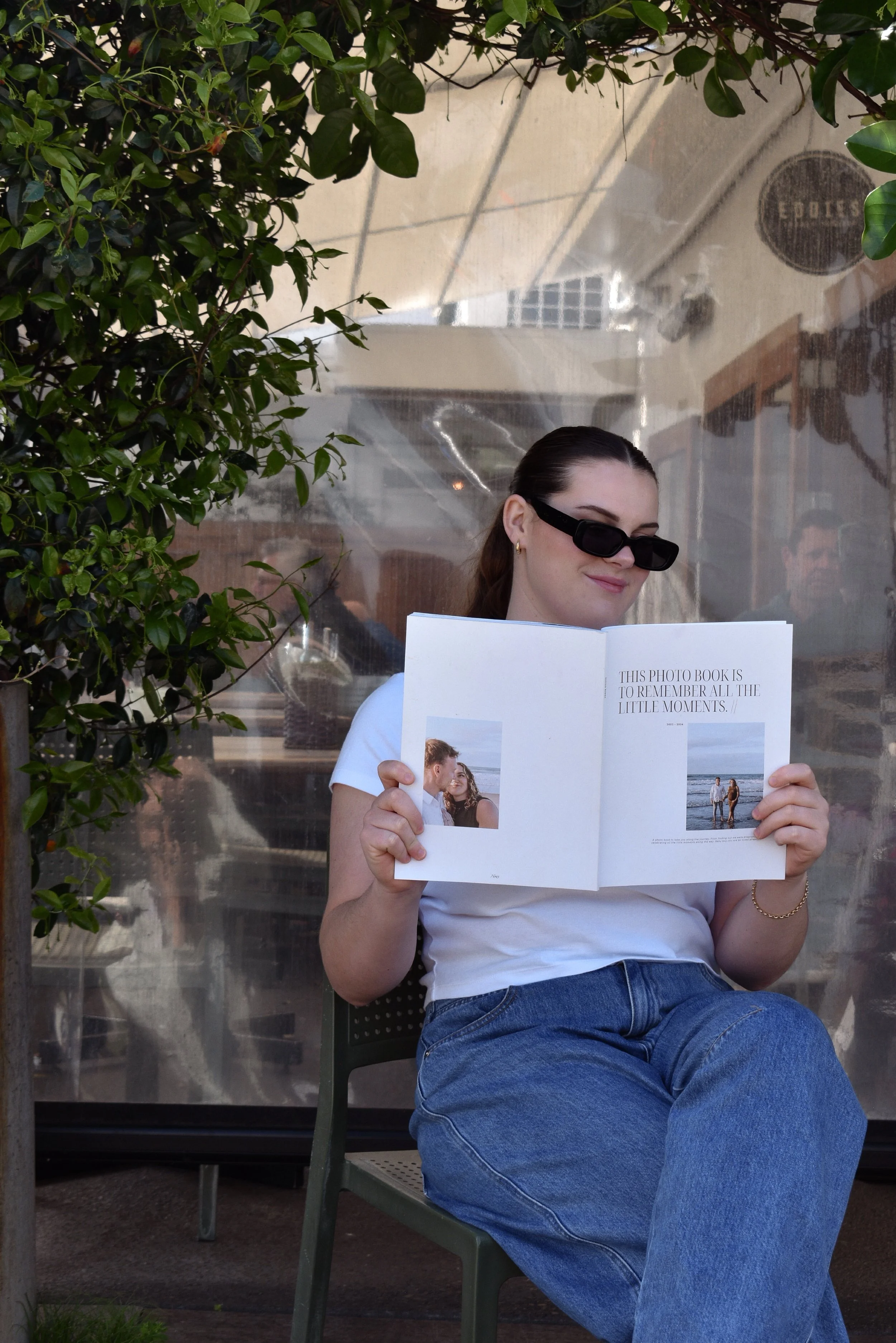 A woman wearing sunglasses and a white t-shirt is sitting outdoors on a green chair, looking at a photo book she is holding. She is surrounded by greenery and a transparent plastic barrier. Inside the book, there is a picture of a couple on the beach and a quote that reads, 'This photo book is to remember all the little moments.'