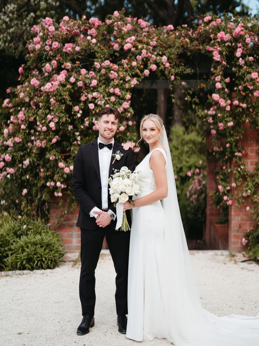 Wedding couple standing outdoors in front of a floral arch, groom in a black tuxedo and bow tie, bride in a white gown holding a bouquet of white flowers.