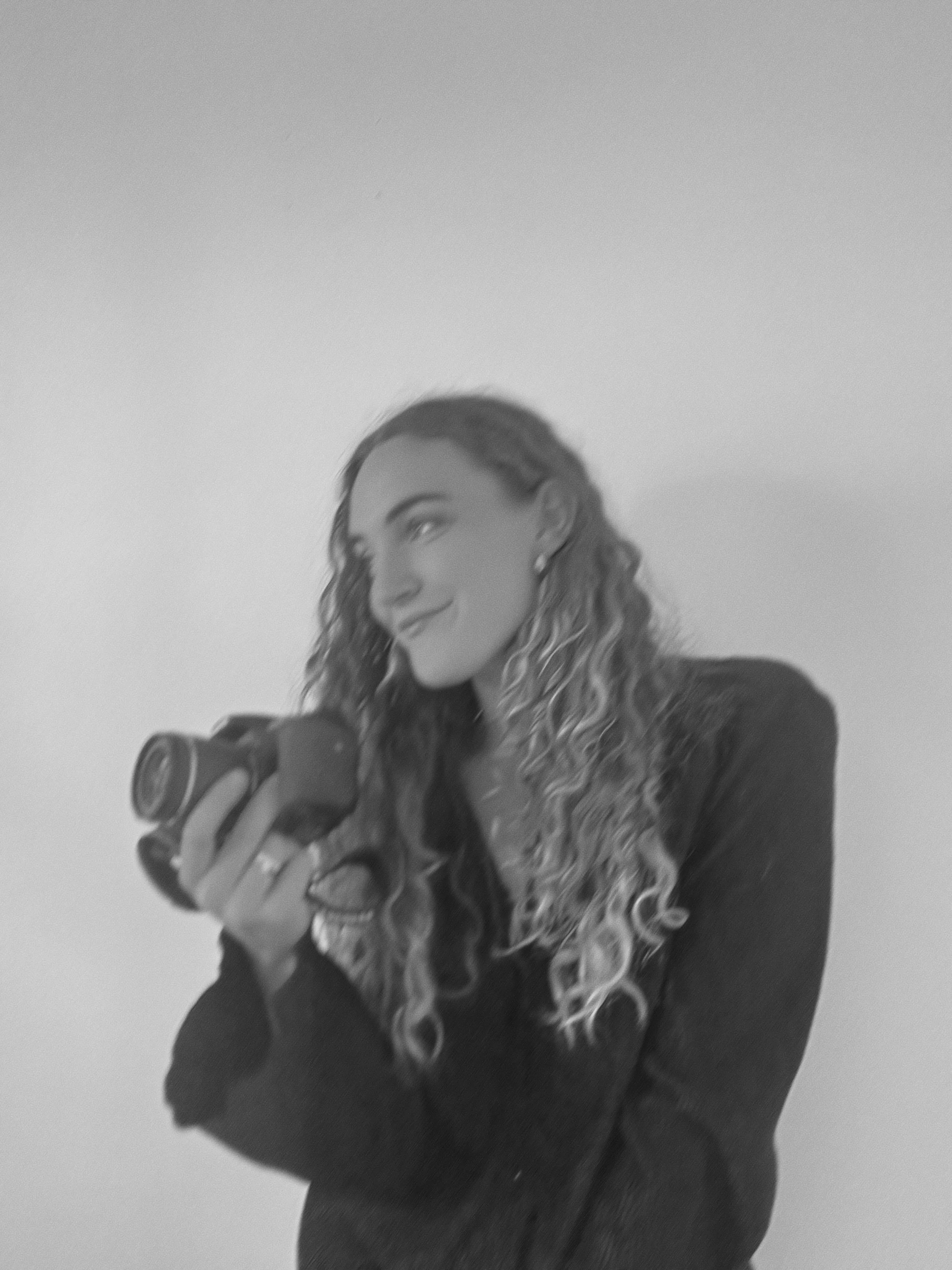 A woman with curly hair holding a camera, smiling, against a plain background.