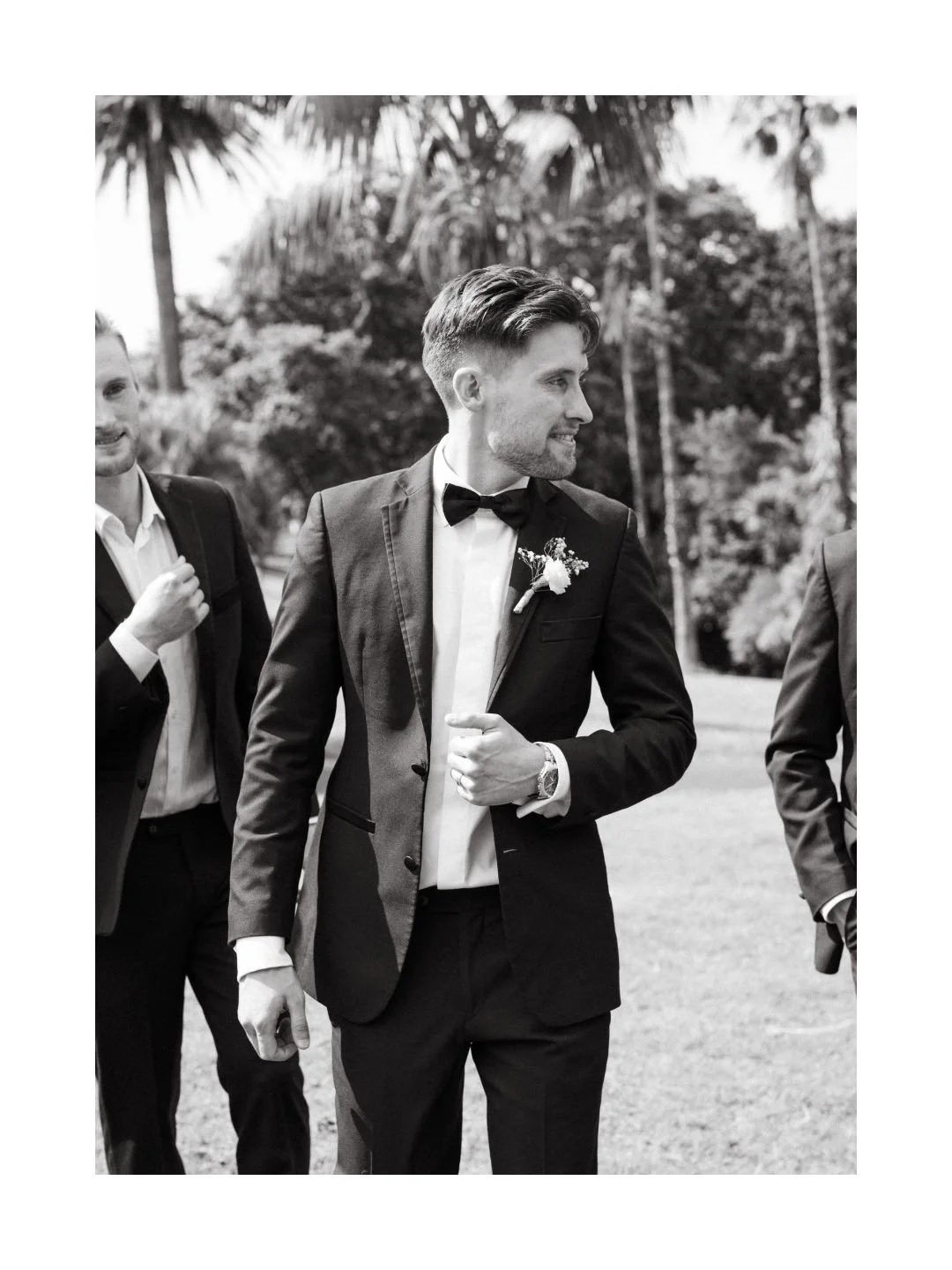 Black and white photo of a man in a tuxedo with a boutonniere, wearing a watch, smiling and looking to the side, walking outdoors with other men dressed in suits, with palm trees in the background.